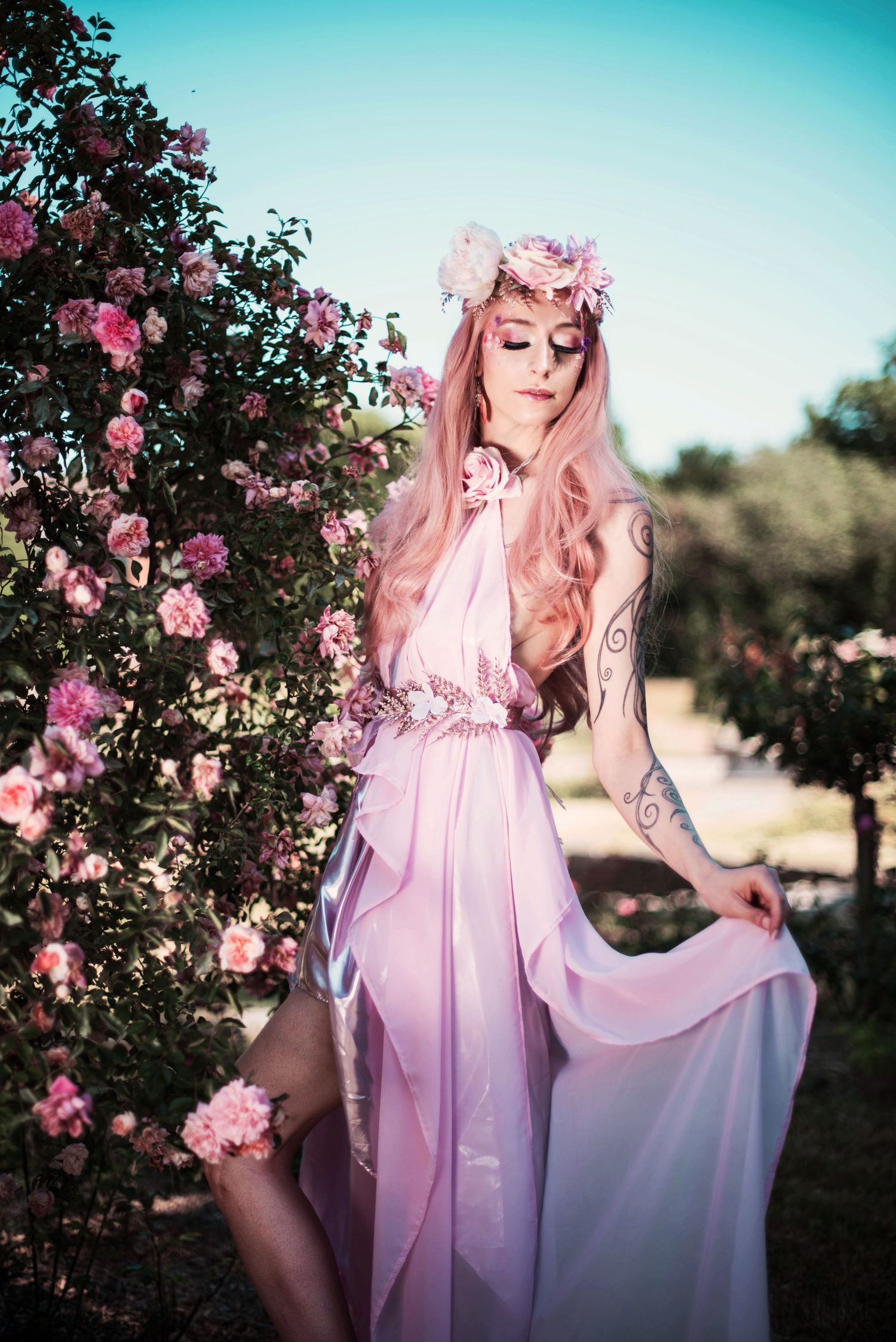 A figure in a flowing pink dress poses among blooming rose bushes at sunset.