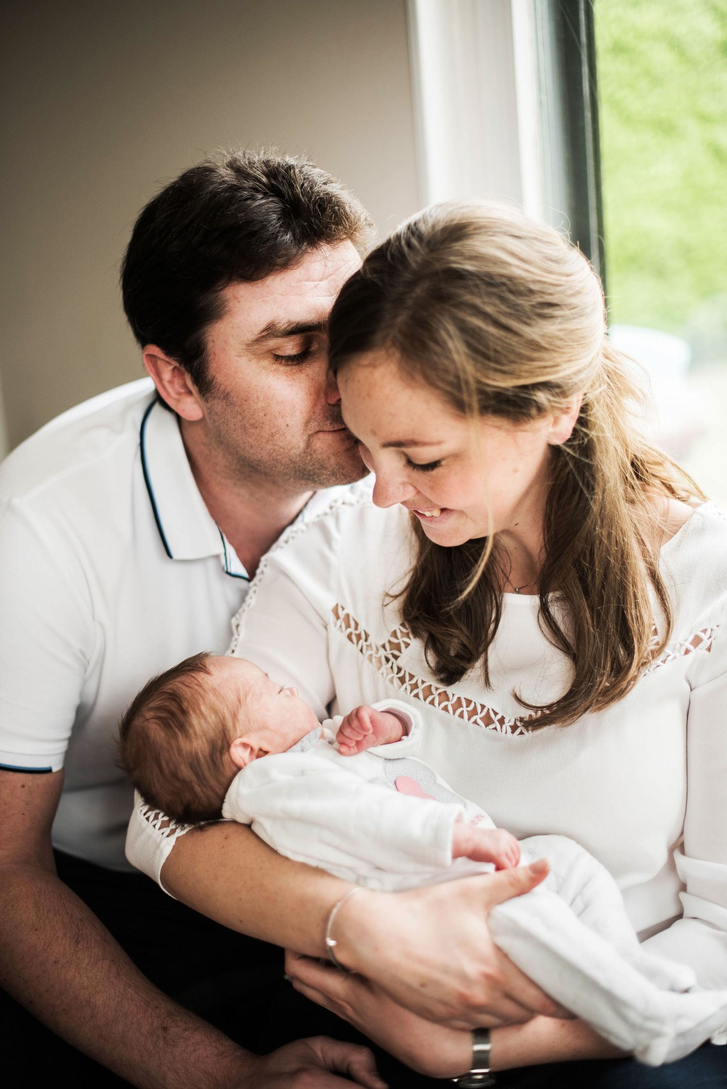 Intimate family moment with parents gazing at newborn by window.