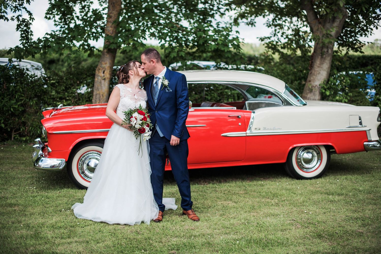 Wedding couple poses with classic red vintage car on green lawn.