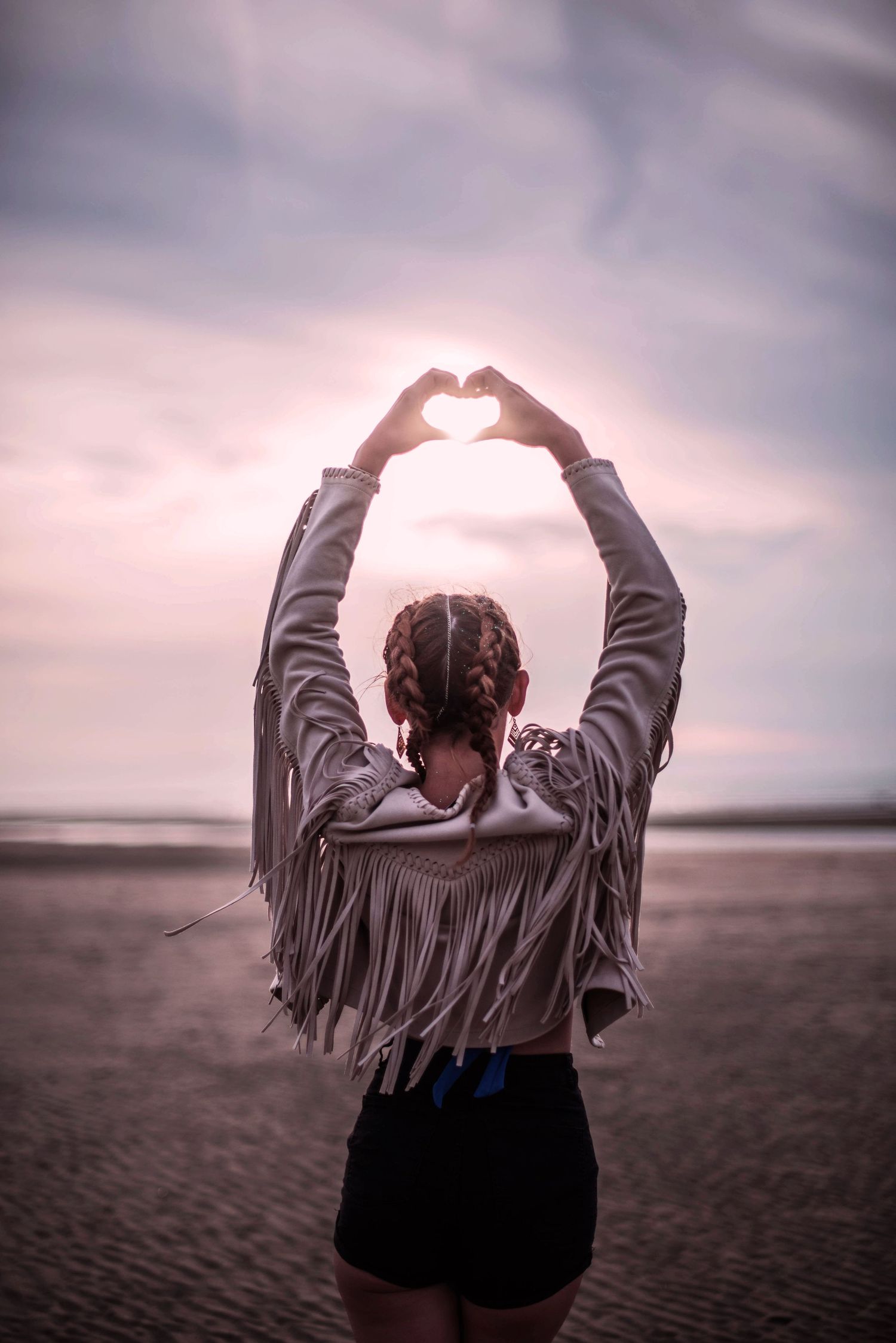 Silhouette making heart shape with hands against sunset sky wearing fringed jacket.