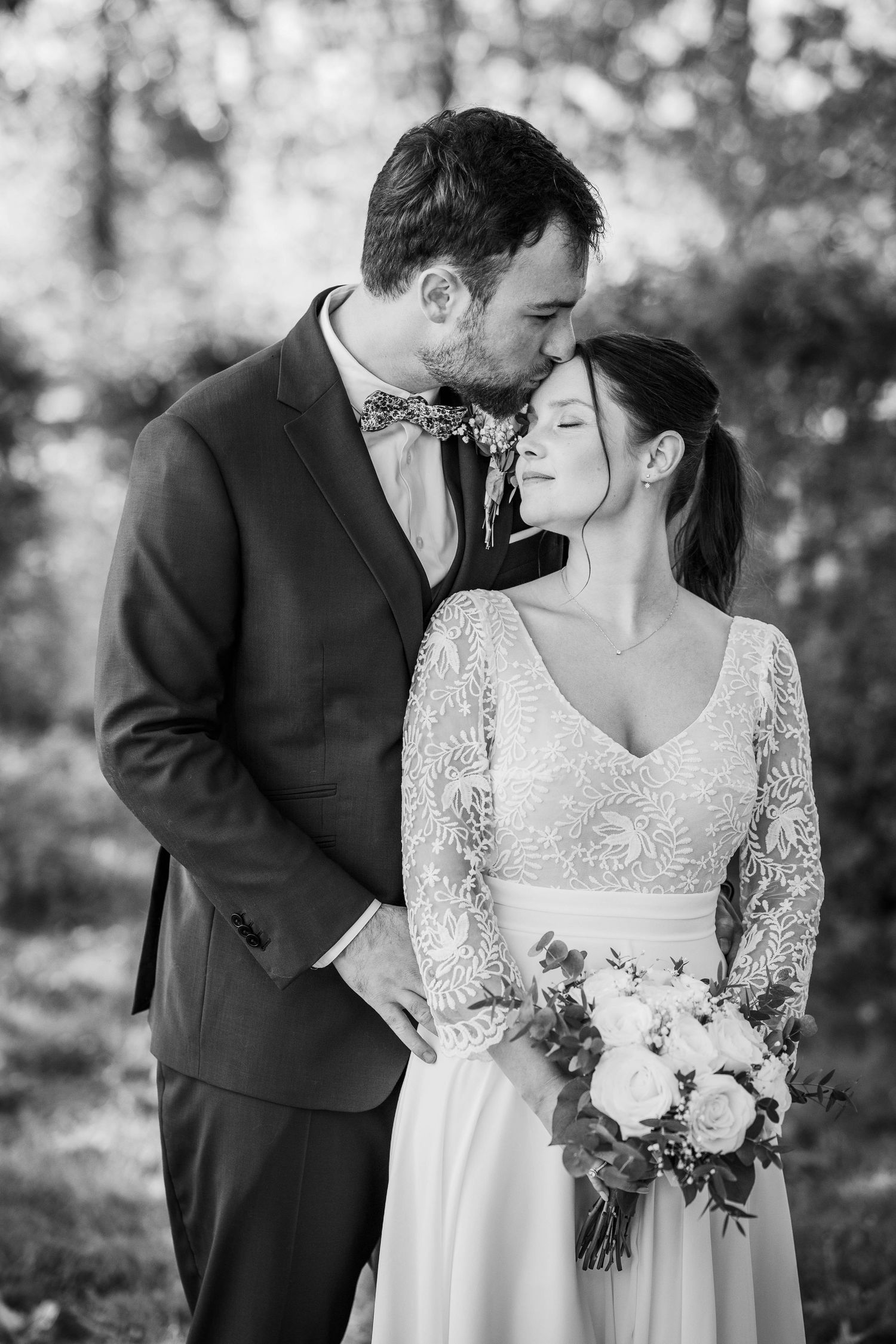 Black and white wedding photo of groom kissing bride on forehead while she holds a bouquet outdoors.