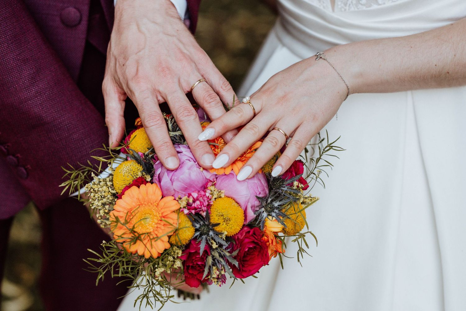 Newlyweds display wedding rings on a vibrant bridal bouquet of orange, pink, and red flowers.