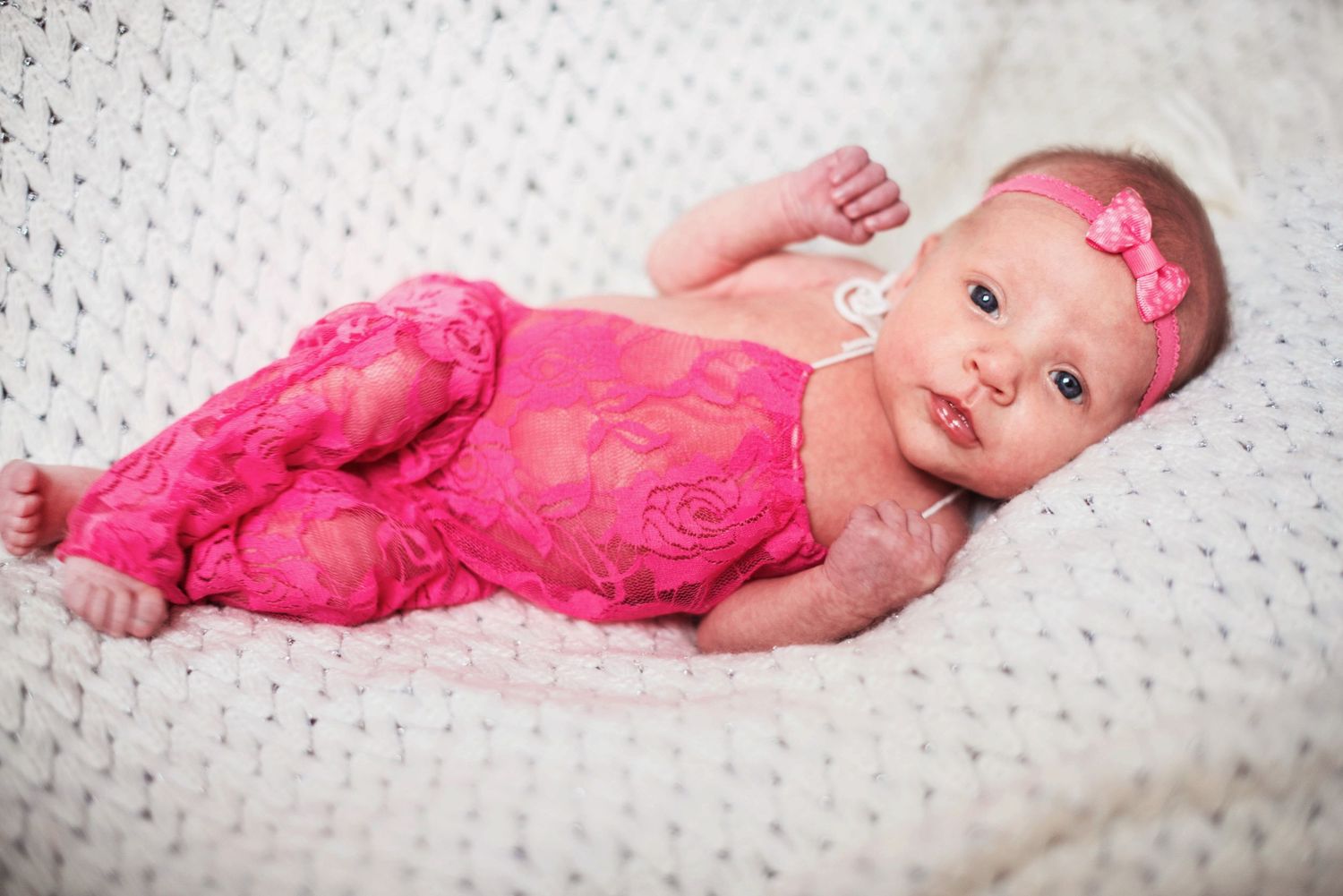 Newborn baby wearing pink lace outfit lying on white textured blanket.