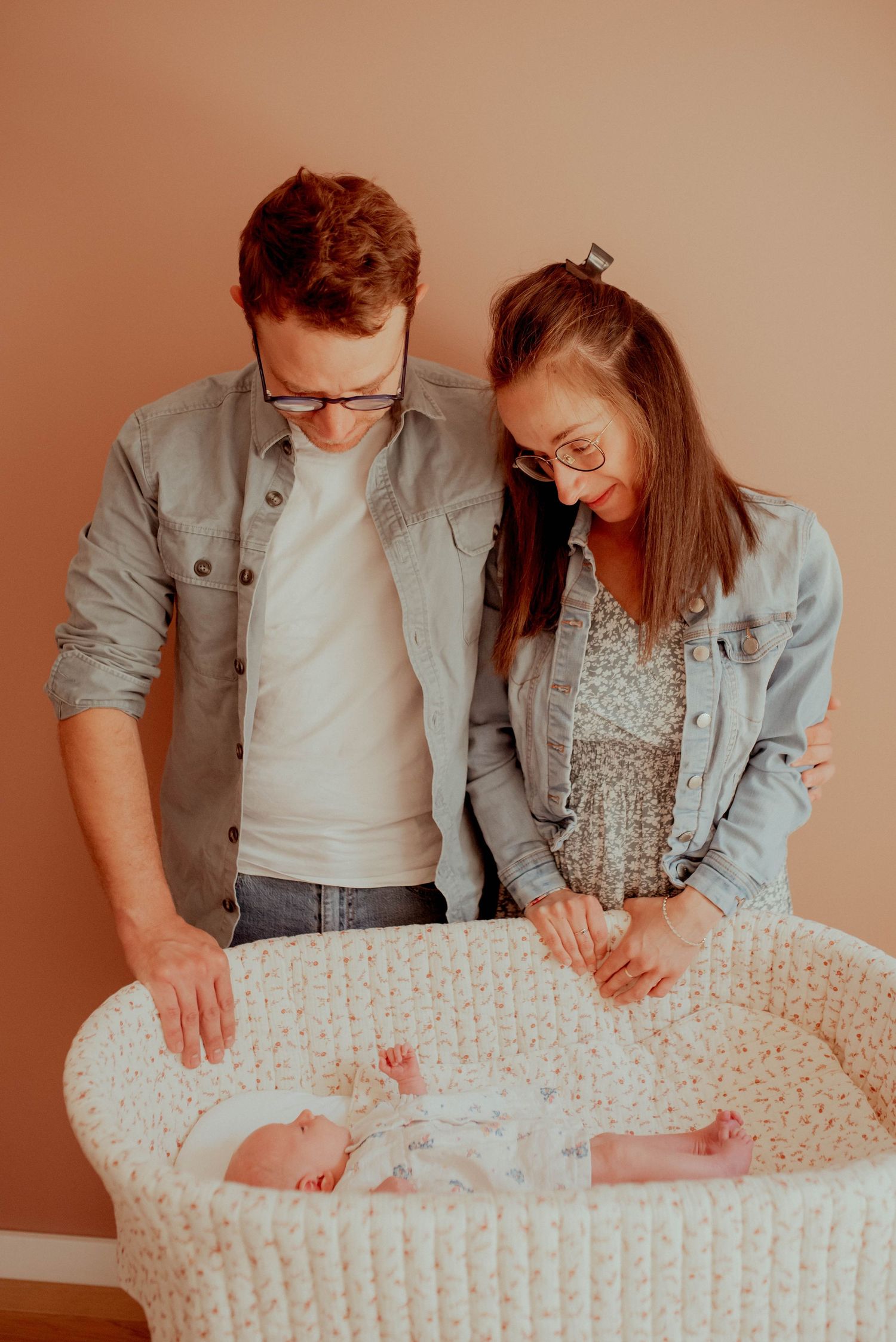 Parents lean over white patterned bassinet to admire their newborn.