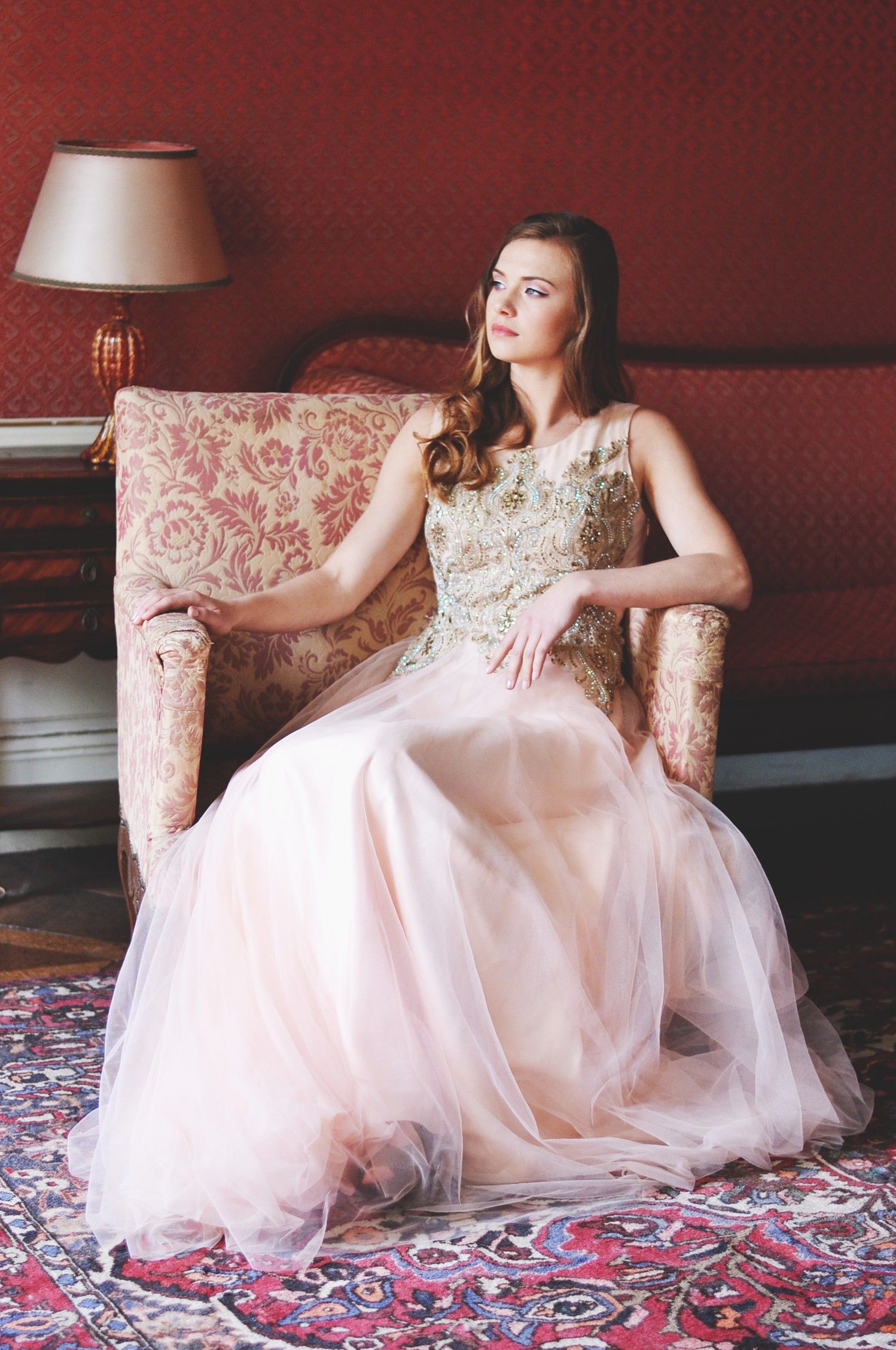 Elegant model in pink tulle gown sits gracefully on vintage floral chair.