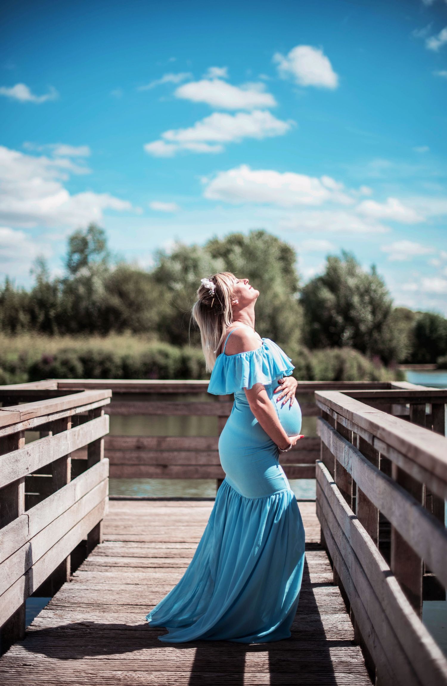 Silhouette of an expectant mother in a flowing blue dress posing on a wooden boardwalk under bright blue skies.