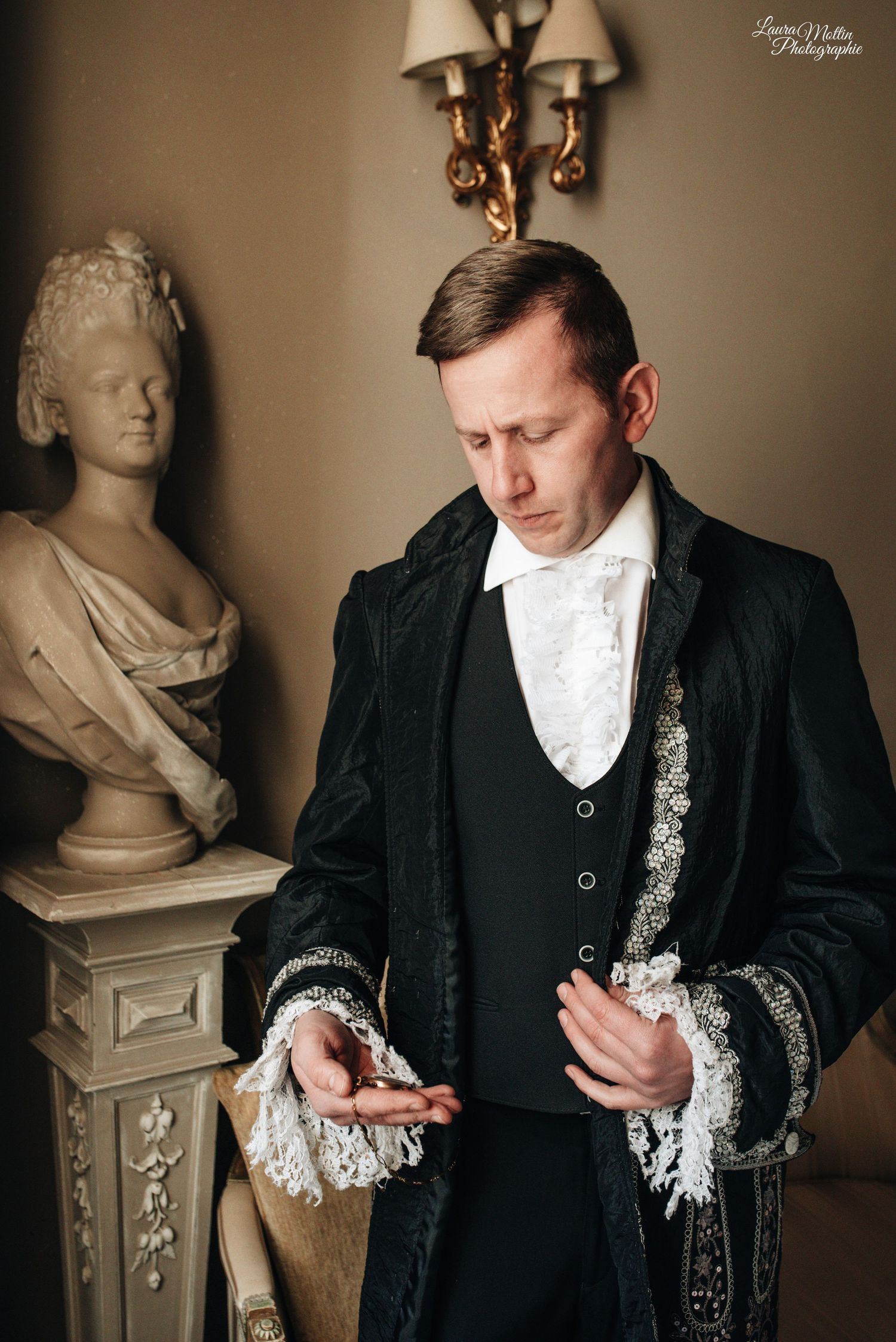 Close-up of formal black suit with white cravat and ornate silver accessories against neutral background.