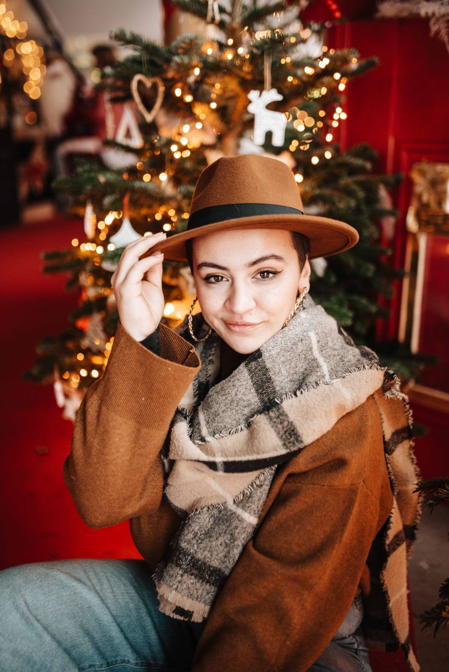 Cozy holiday portraits with brown hat and plaid scarf against Christmas tree lights.