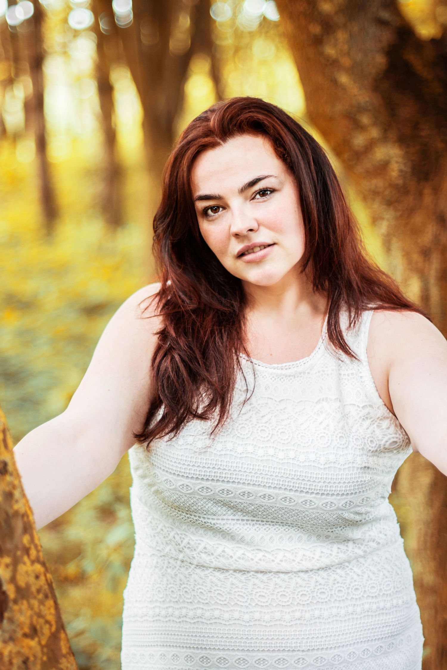 Woman in white lace dress poses against golden sunset in woods.