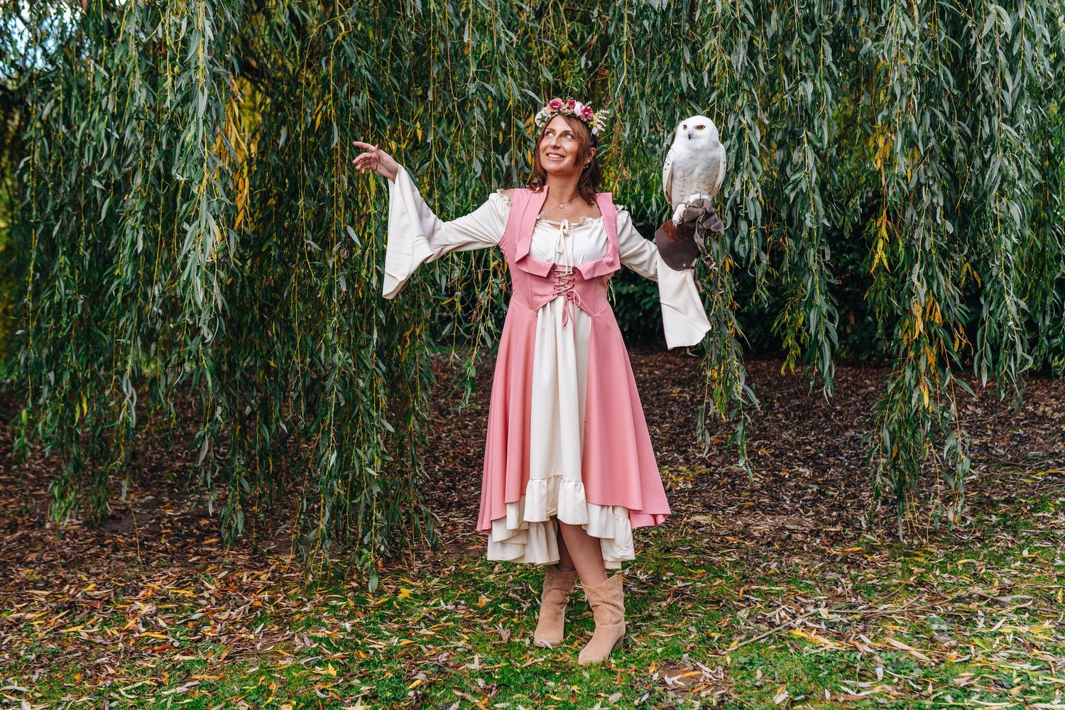 Woman in pink dress twirls playfully under weeping willow trees in garden setting.