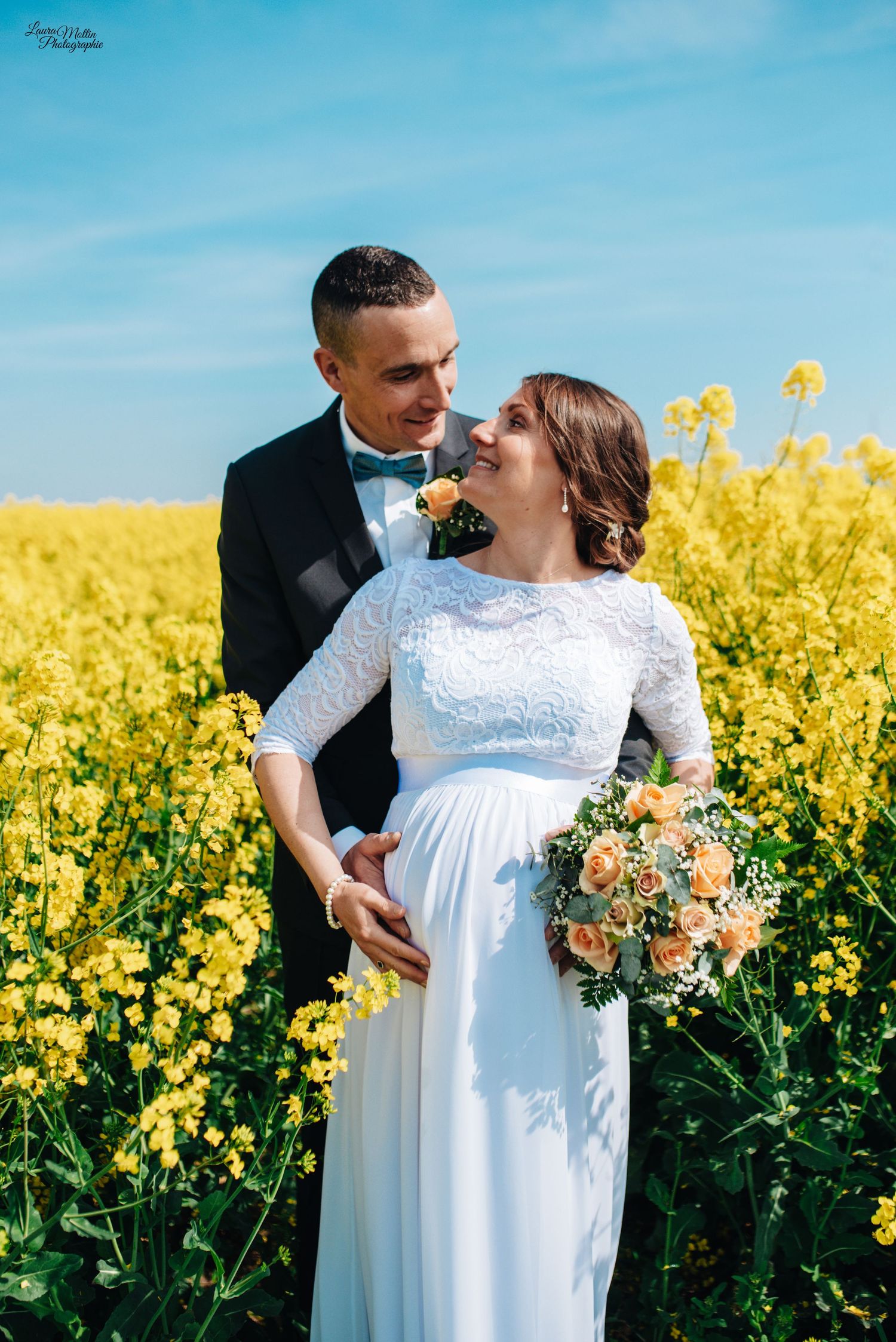 Wedding couple poses intimately in a bright yellow canola field under blue sky.