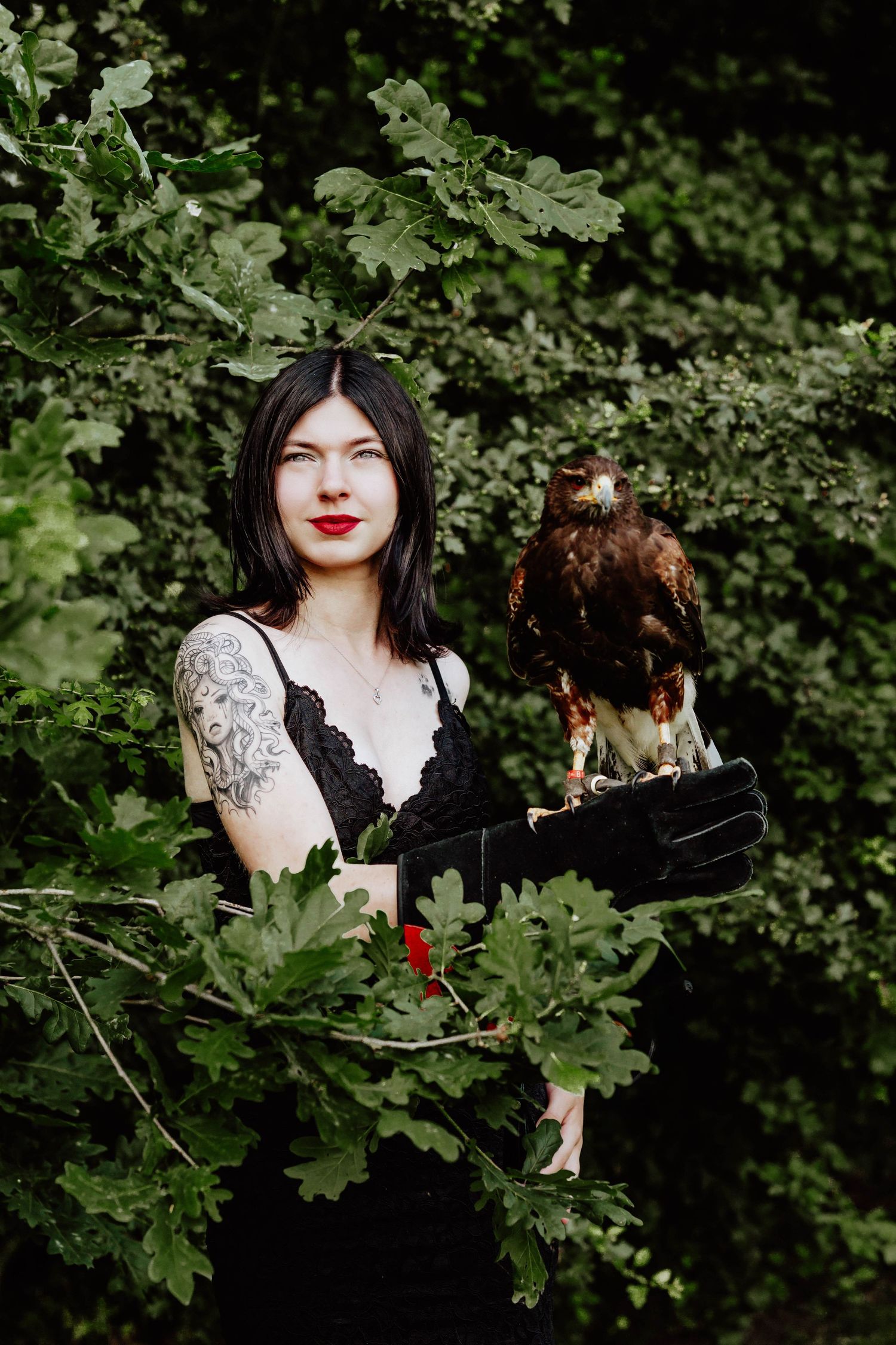 A tattooed woman with black hair holds a hawk on her gloved hand, standing among lush green oak tree branches.