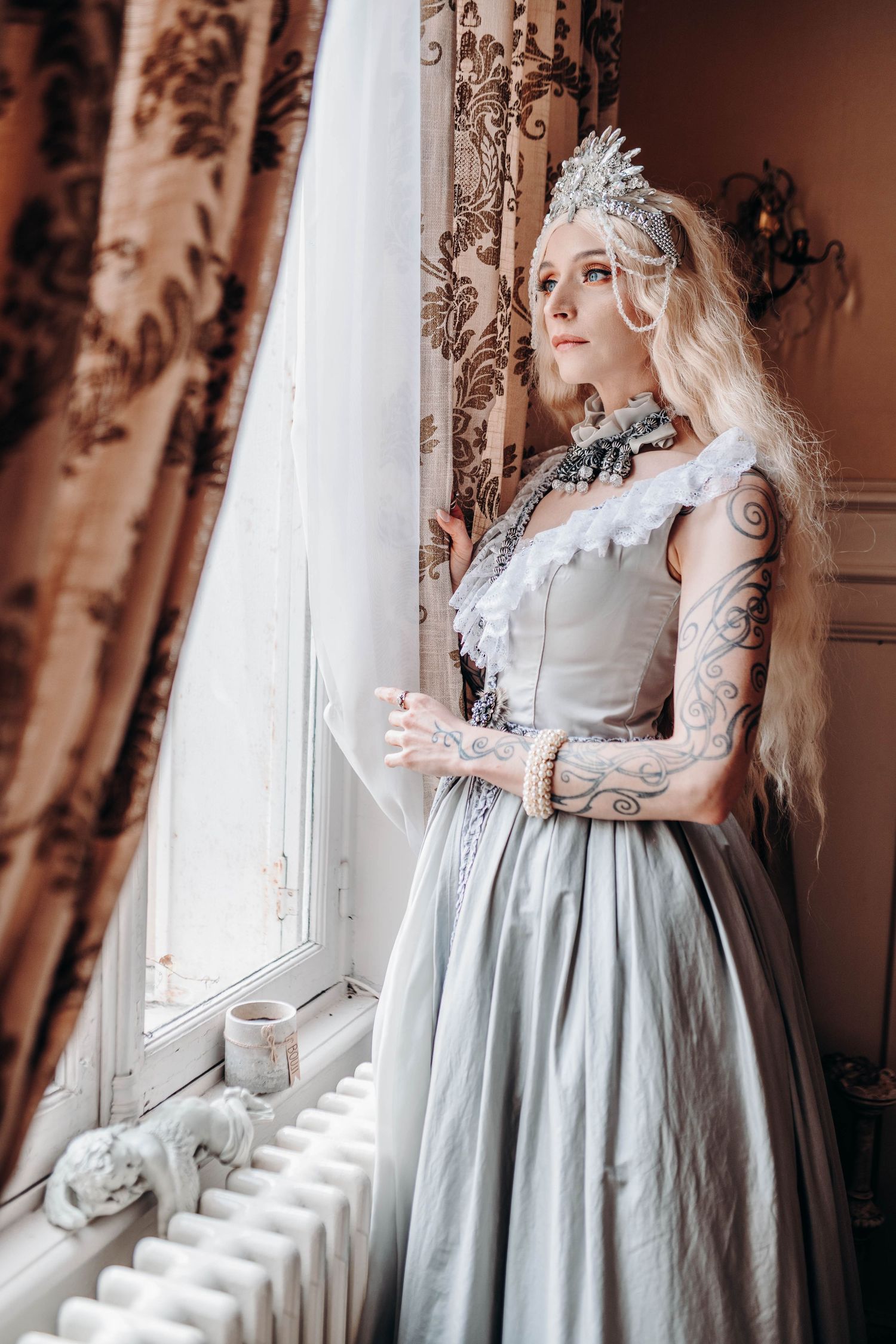 A tattooed blonde woman in a gray Victorian gown and silver crown stands gracefully by a ornate window.