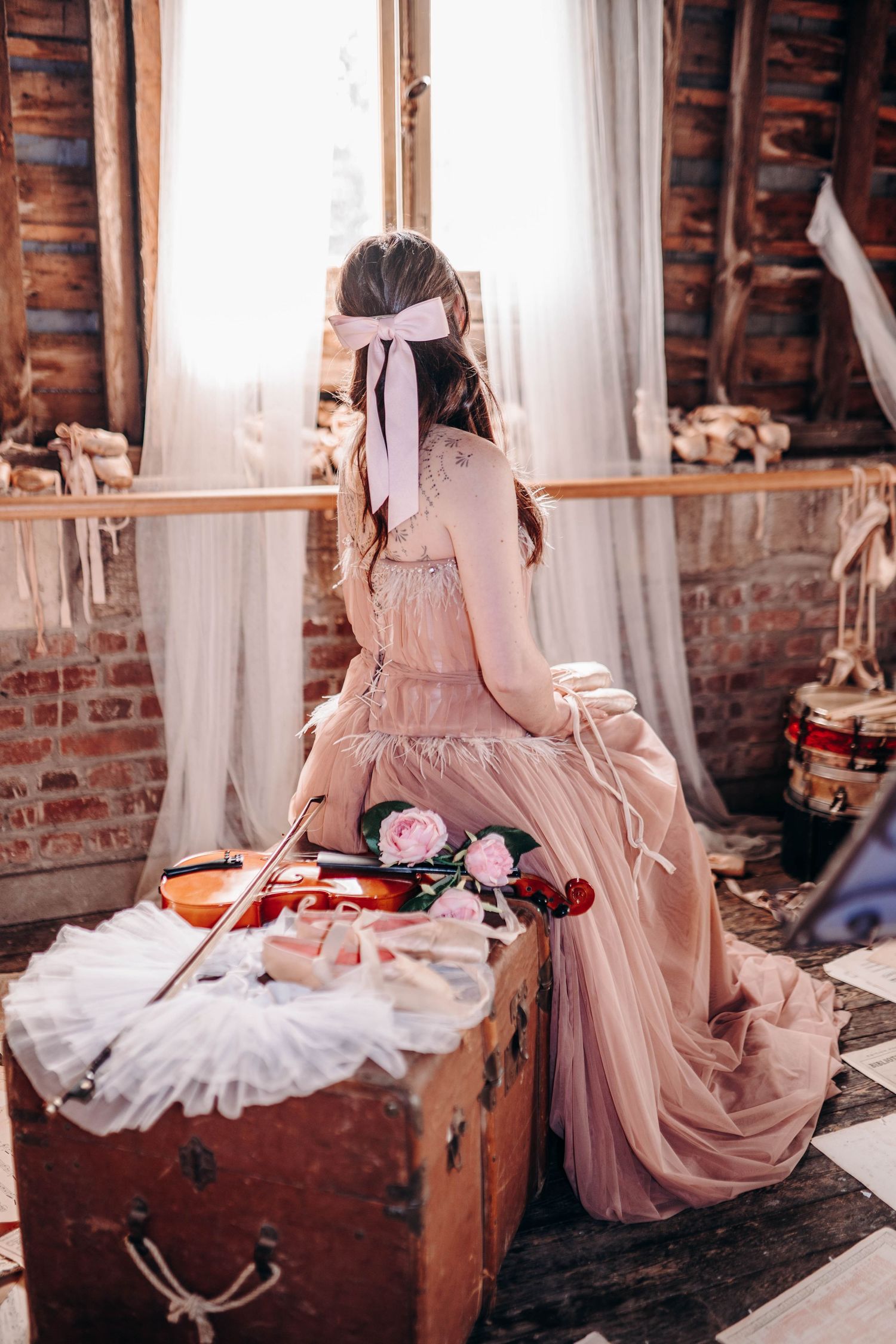Young woman in pink gown sits by window with violin and roses on vintage trunk in rustic attic setting.