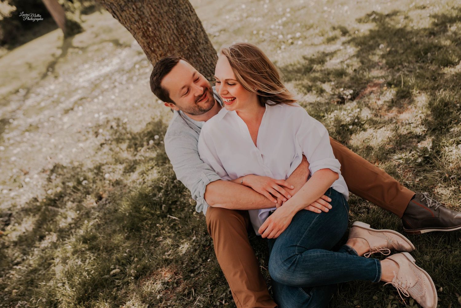 Couple laughing together while sitting on grass during golden hour outdoor photo session.