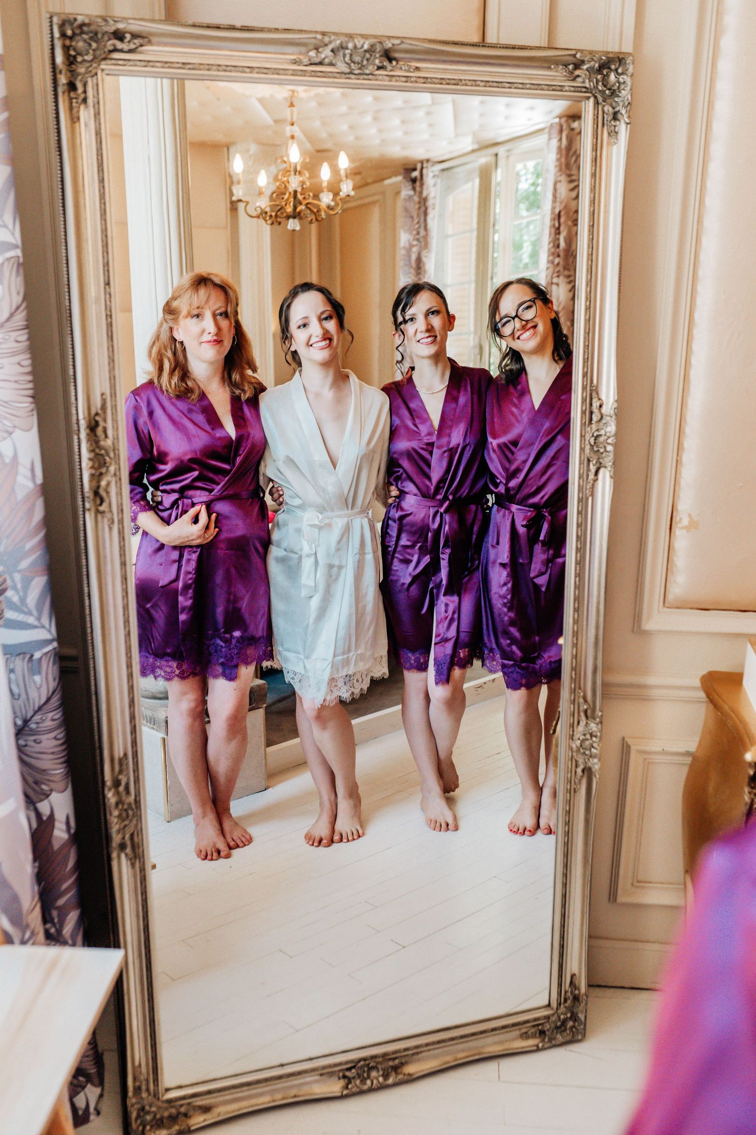 Bride and bridesmaids in purple and white satin robes smiling together reflected in an ornate gold mirror.