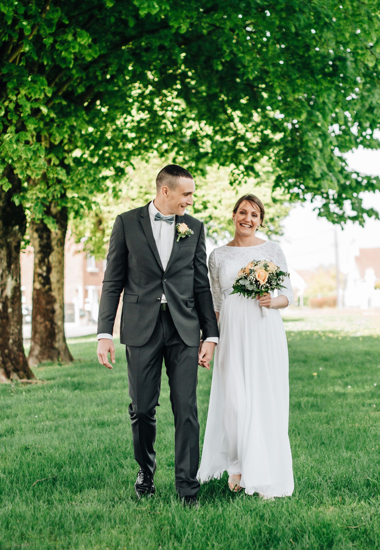 Wedding couple walking together under green trees on manicured lawn.