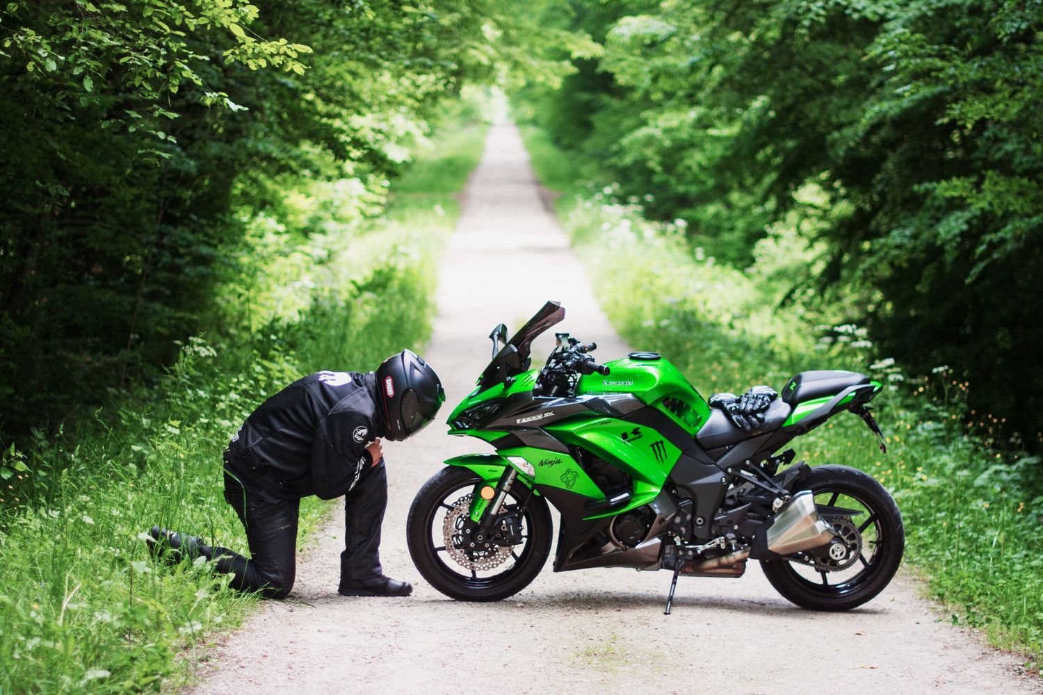 Motorcyclist inspecting bright green sport bike on tree-lined path.