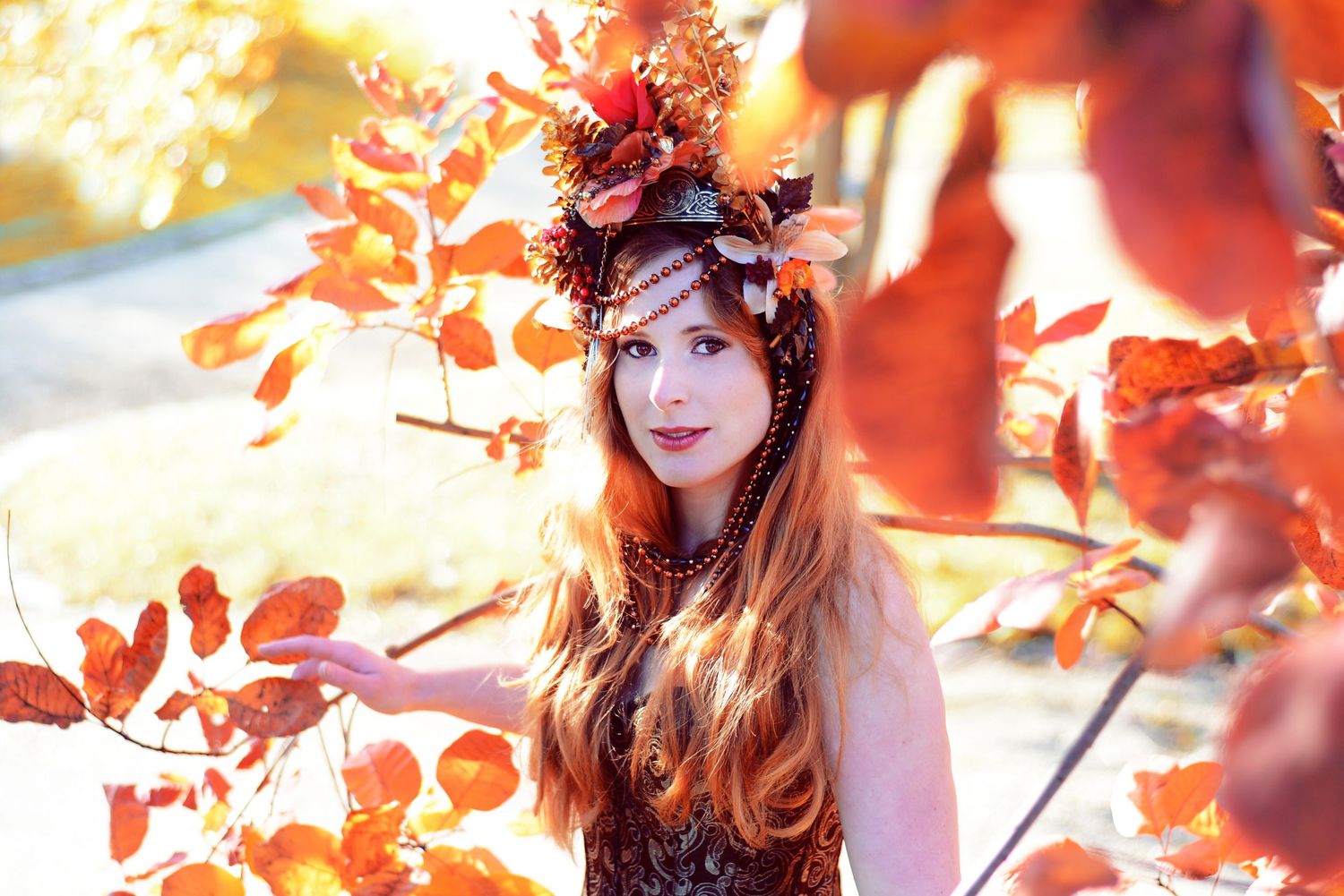 Person wearing autumn-themed headpiece posing among red fall leaves in sunlight.