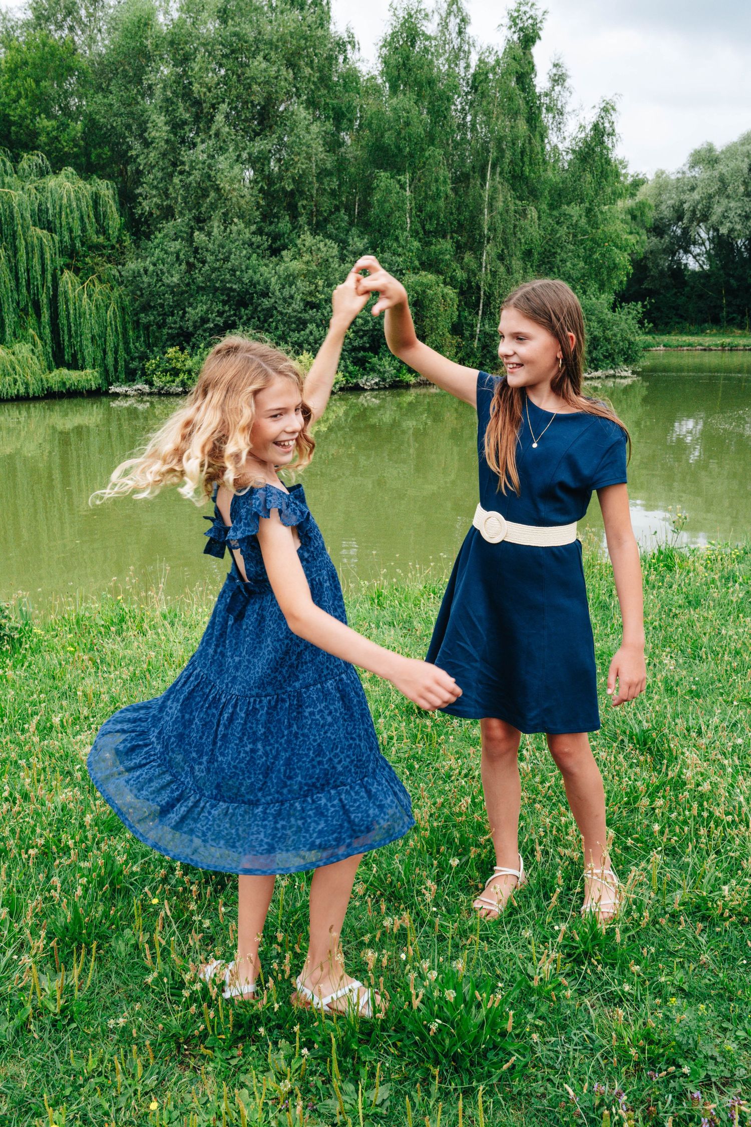 Two children in blue dresses dancing and twirling in a grassy field by a pond.