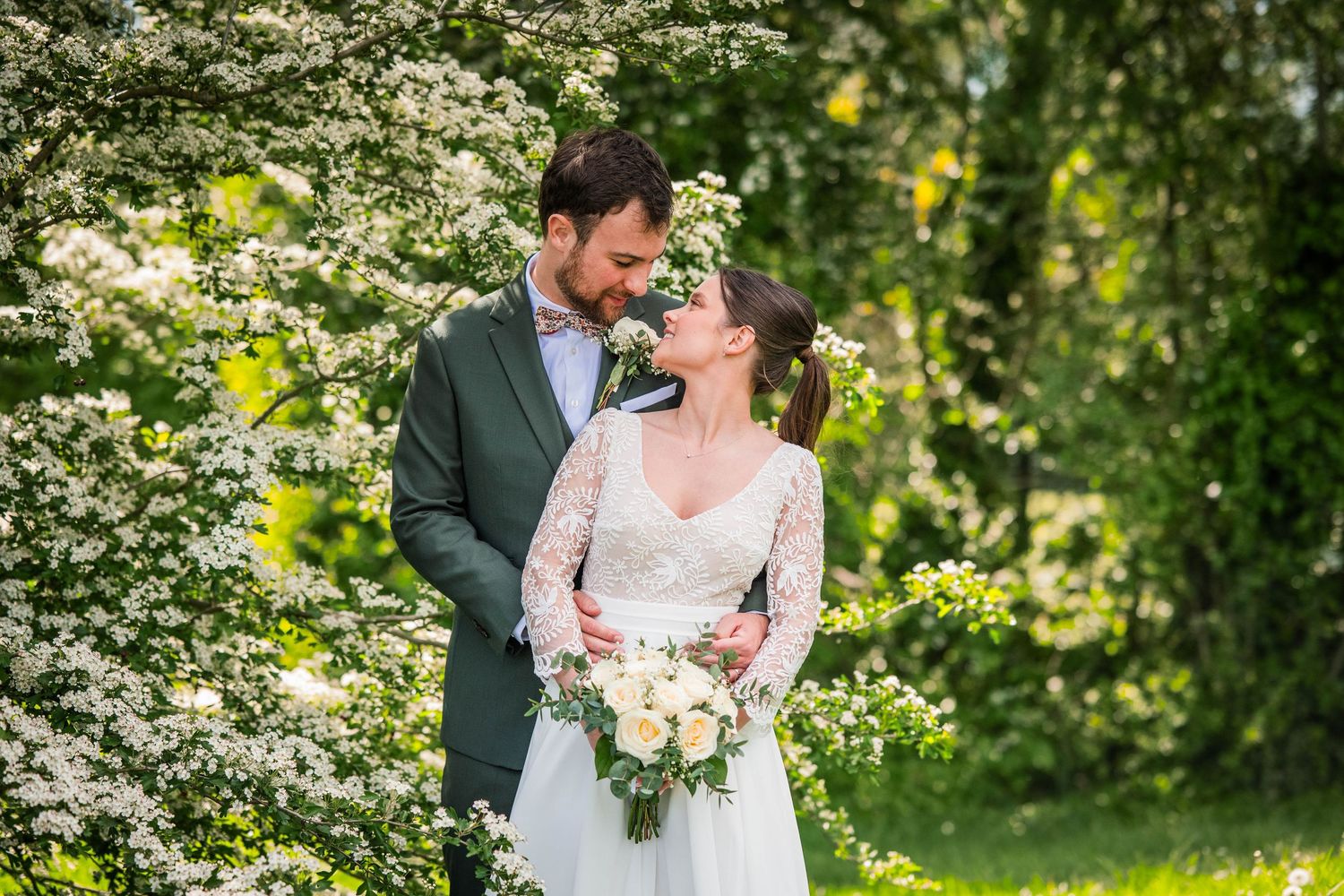 Bride and groom share a romantic moment surrounded by blooming white flowers in a sunlit garden.
