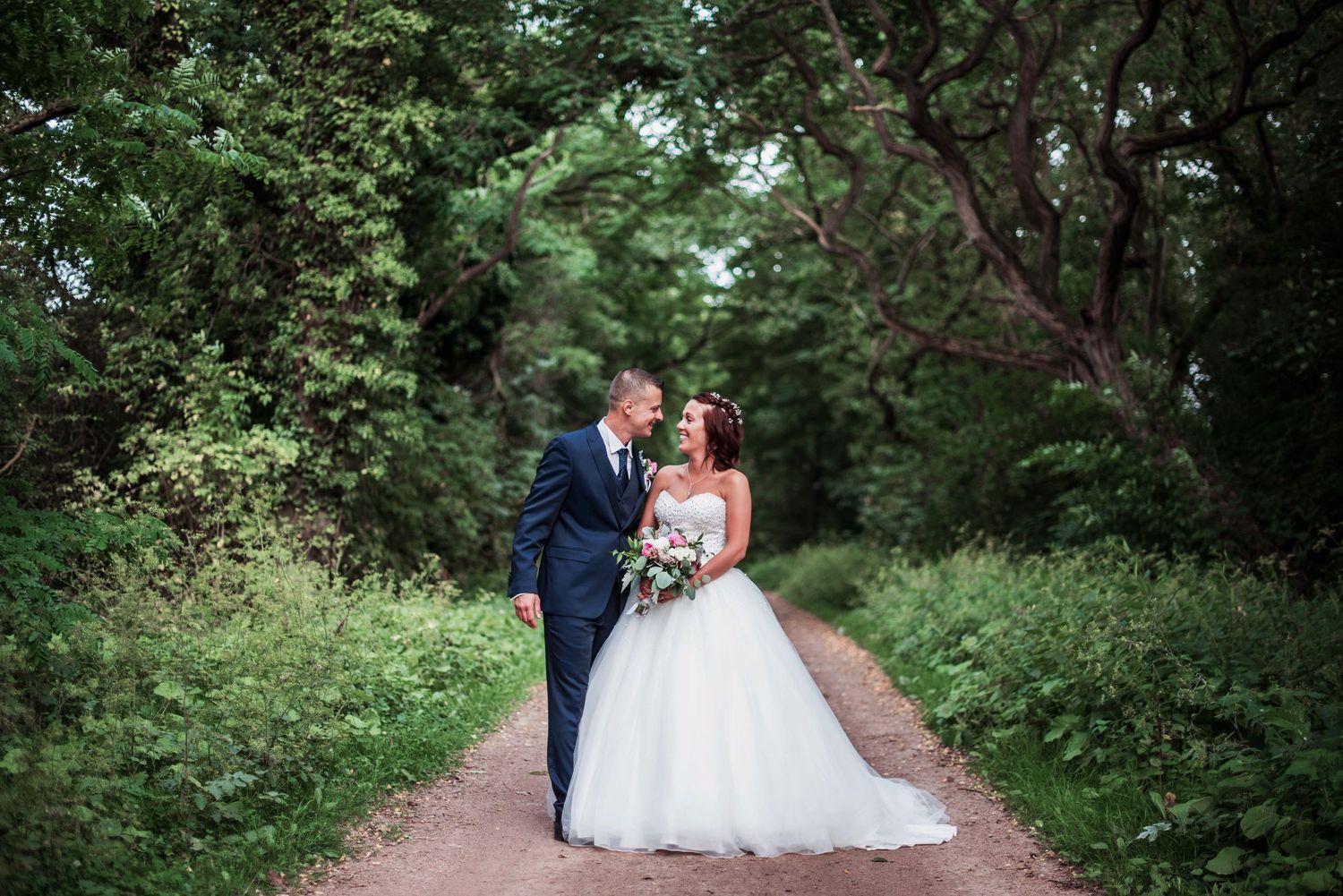 Newlyweds pose on a tree-lined path during their wedding photoshoot.