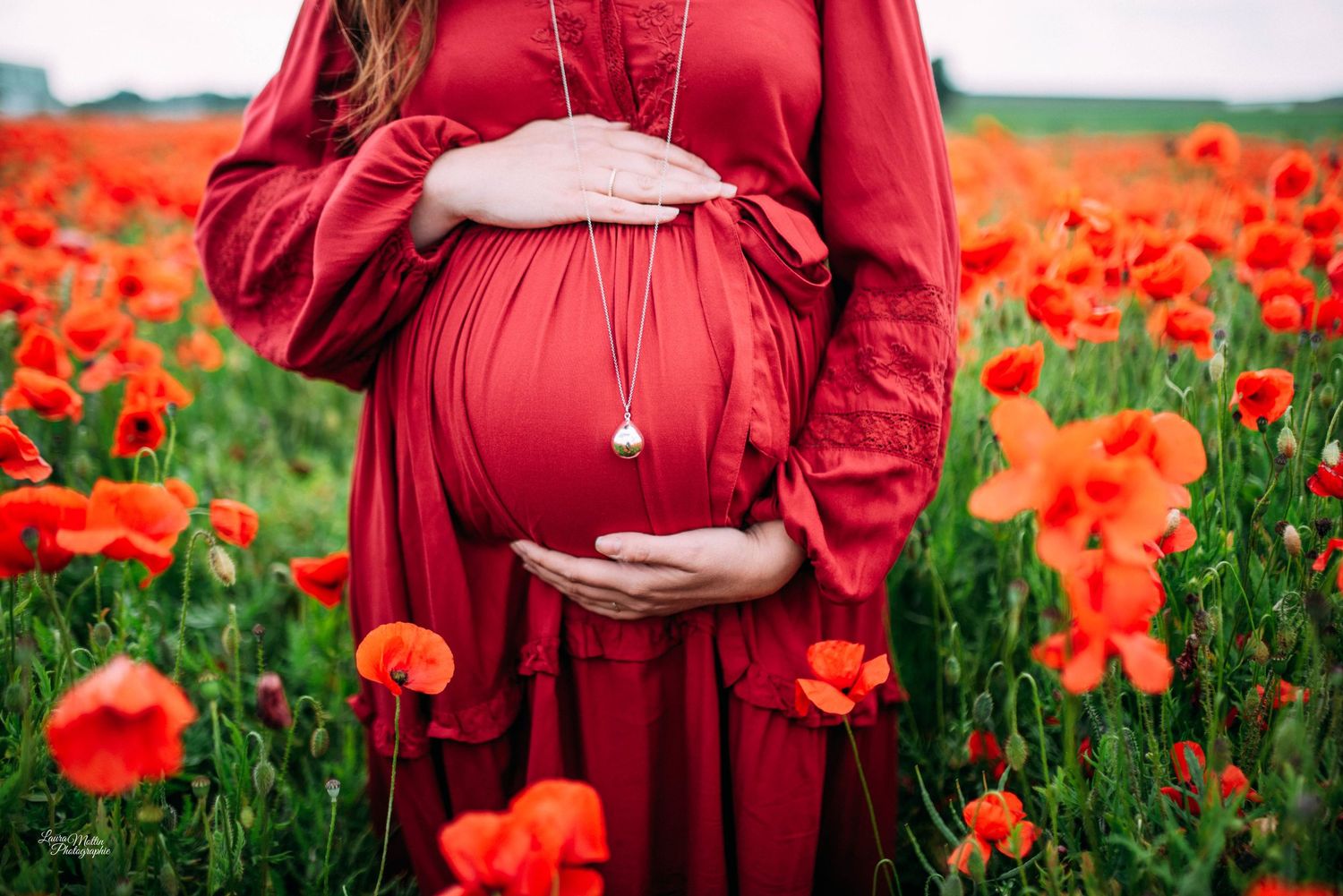 Maternity photo in red dress standing among vibrant orange poppies in a field.