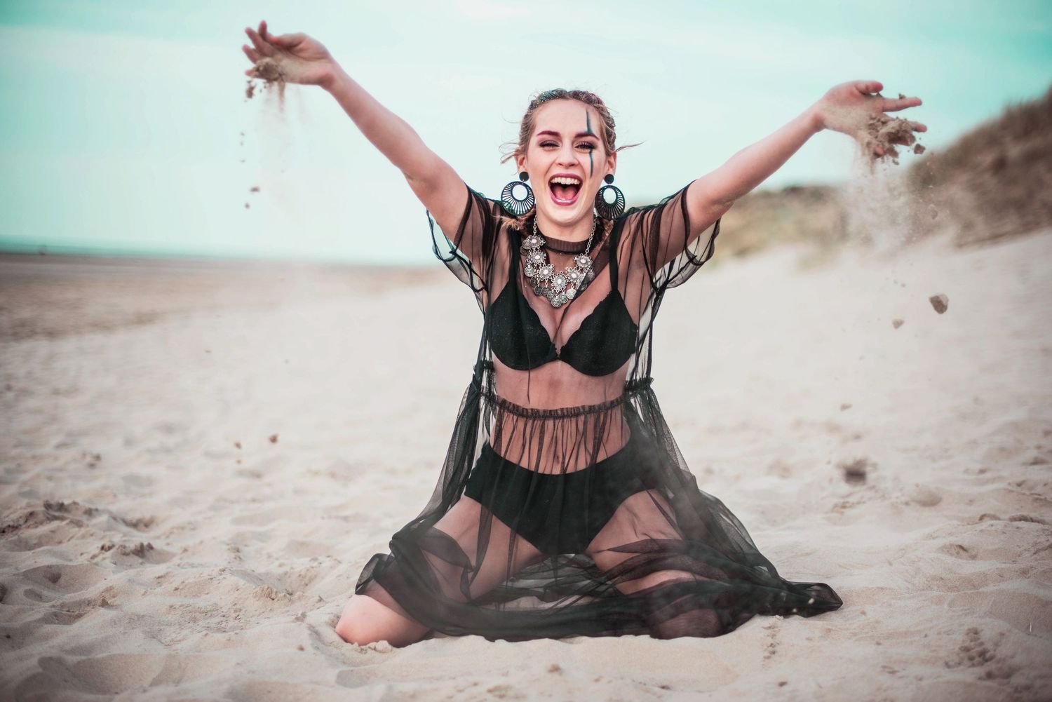 Joyful model in black dress poses expressively on beach during sunset photoshoot.