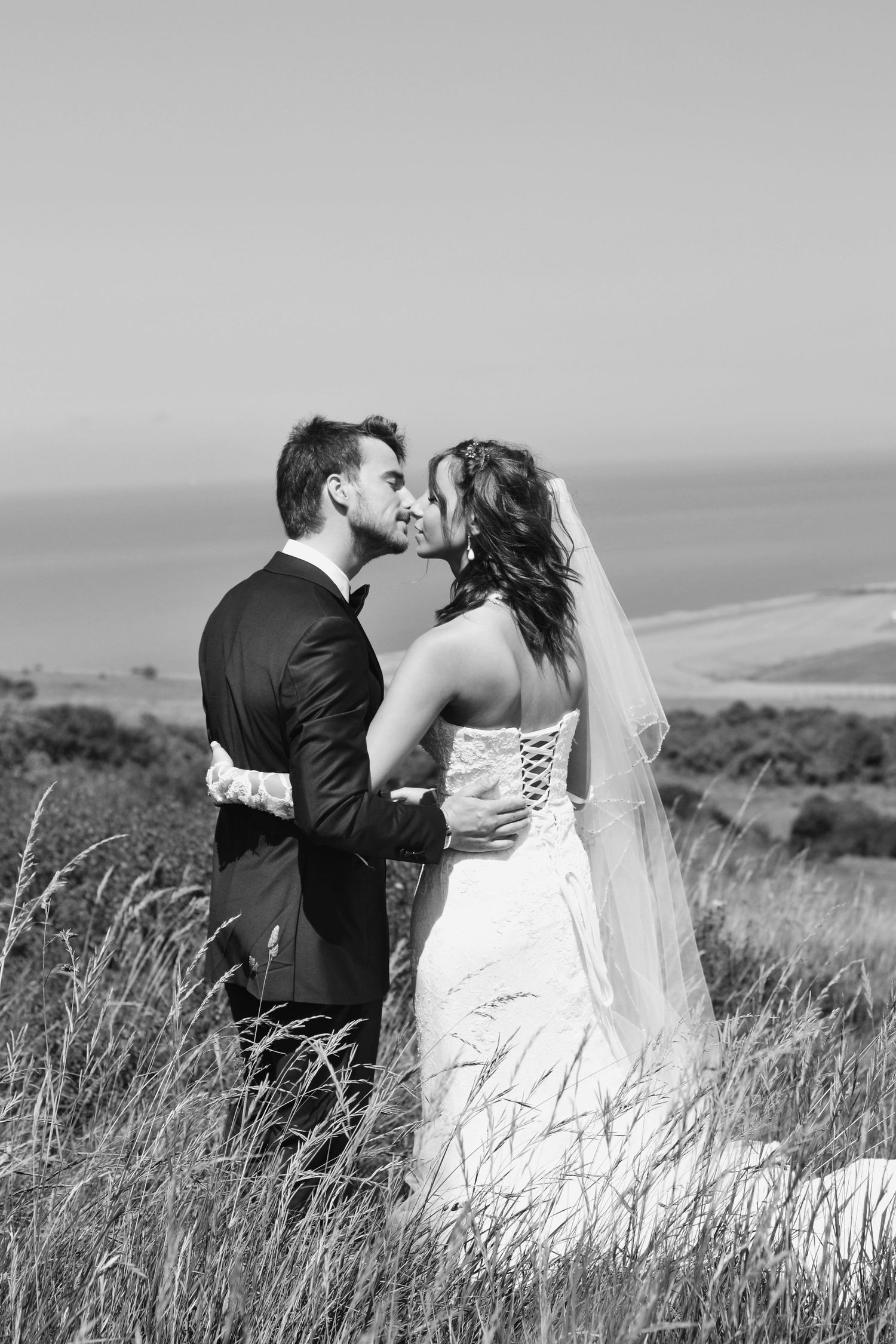 Black and white wedding portrait of couple kissing in tall grass with ocean view.