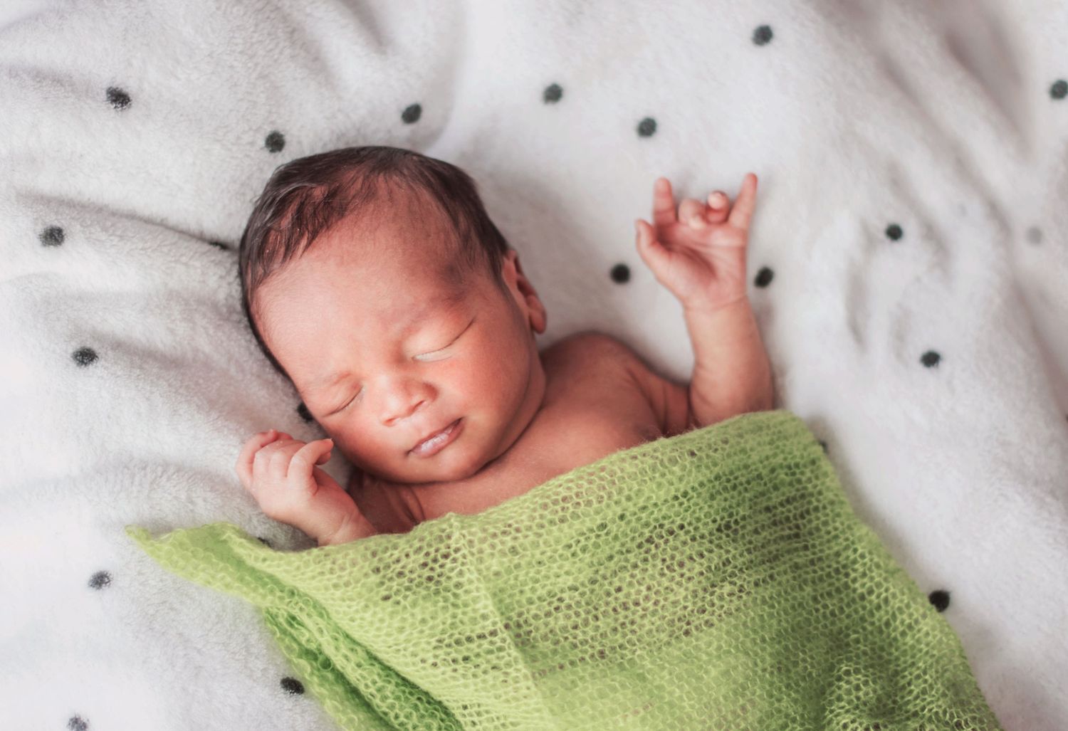 Sleeping newborn wrapped in soft green knit blanket on polka dot bedding.