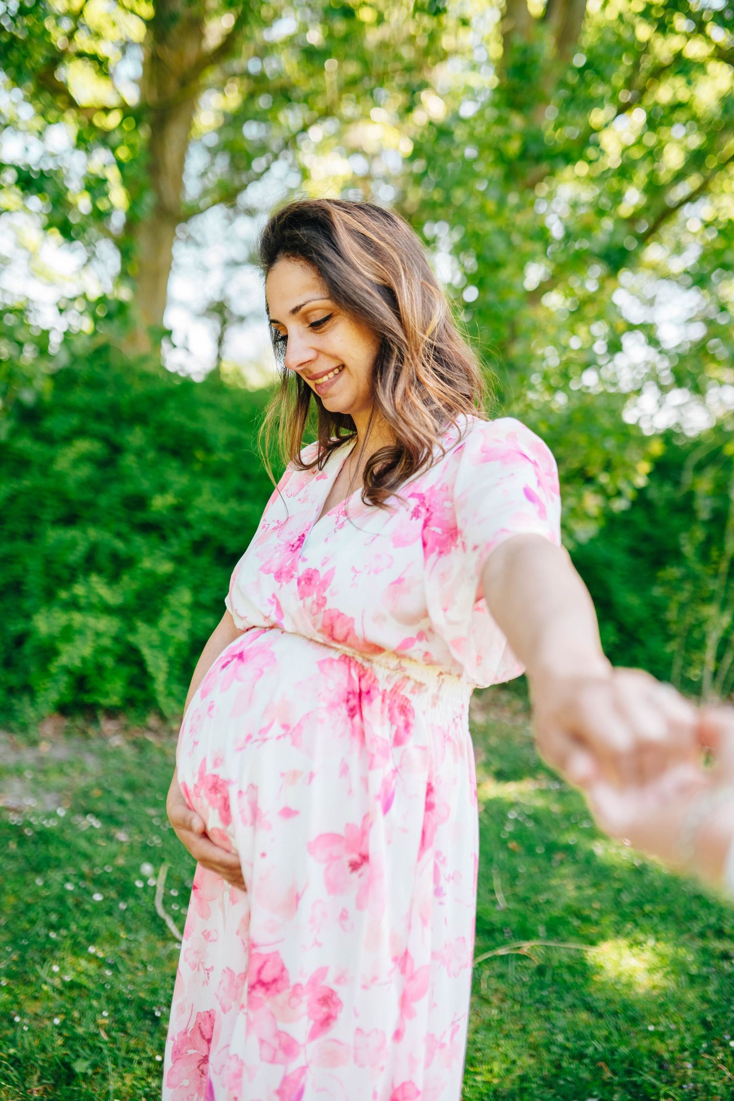 Smiling pregnant woman in pink floral dress holding hands outdoors in lush green park during maternity photoshoot.