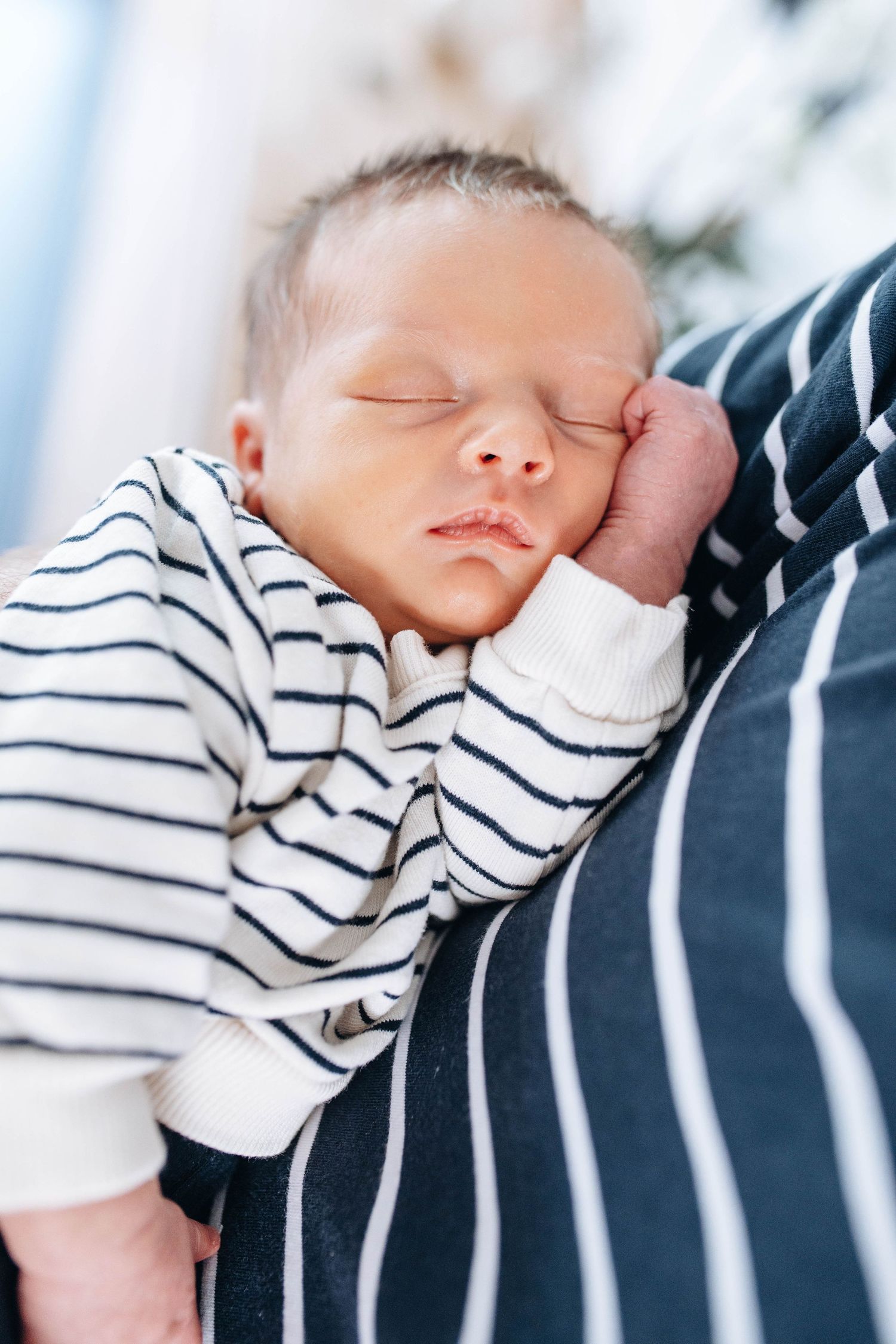 Peaceful newborn baby sleeping in striped onesie, resting cheek on tiny fist against parent's striped shirt.