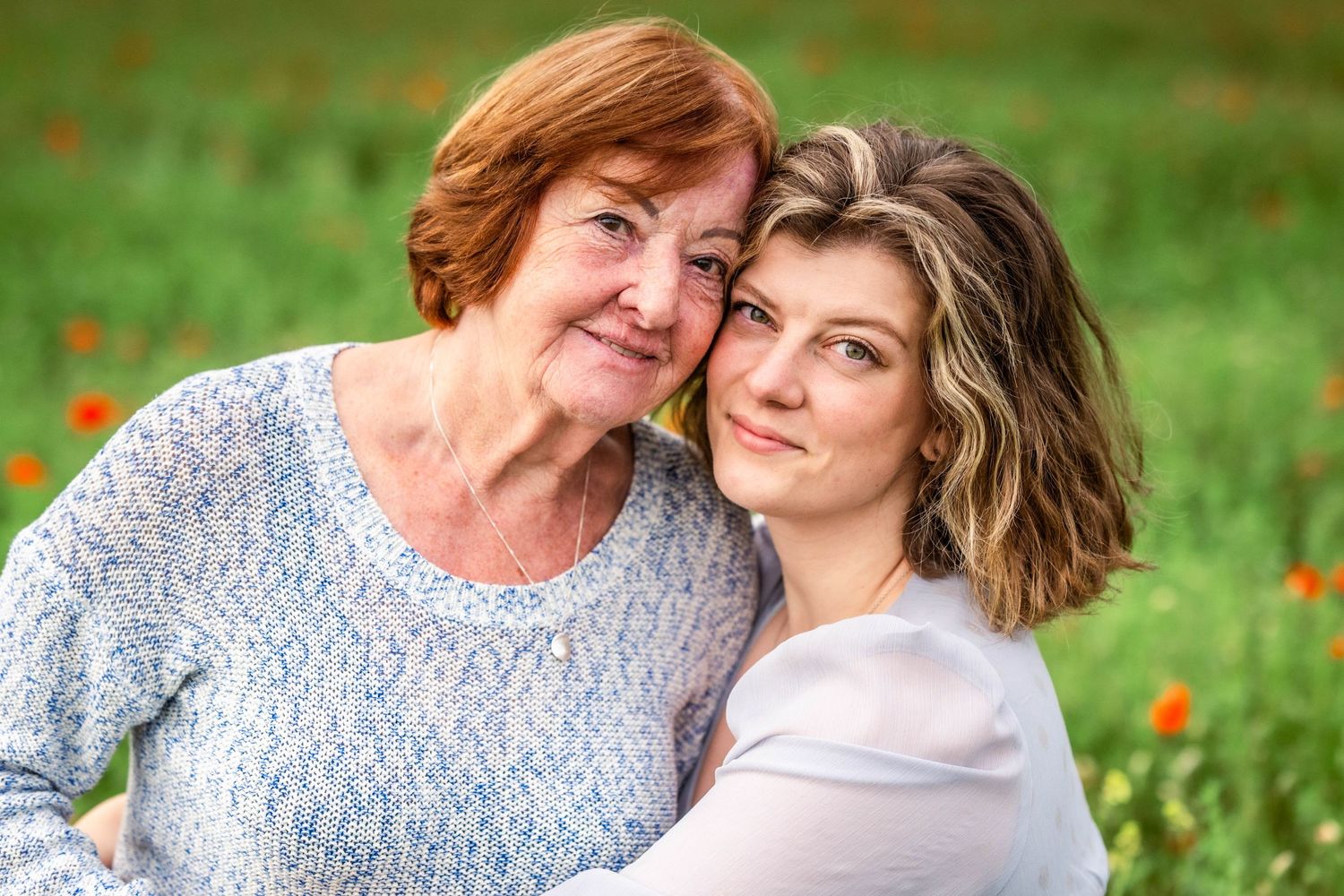 Two women, possibly mother and daughter, share a tender cheek-to-cheek moment outdoors in a green field with red flowers.