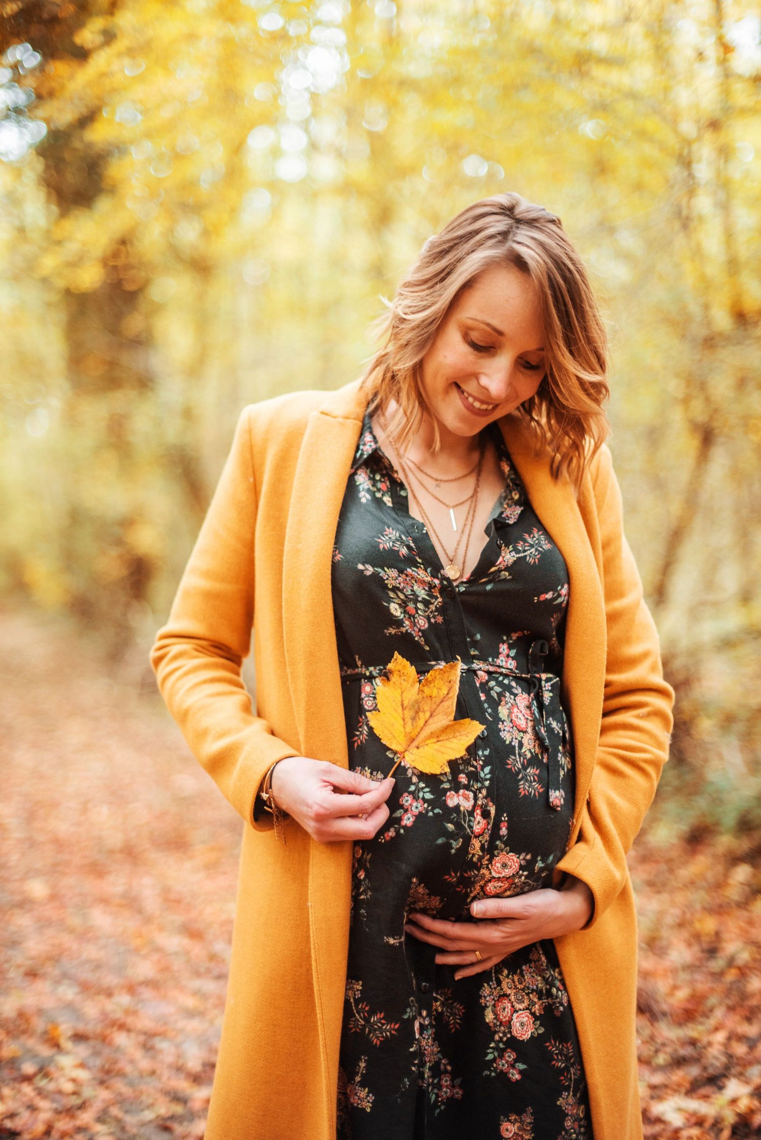 A person in a yellow coat holds an autumn leaf while wearing a floral dress in a fall forest setting.