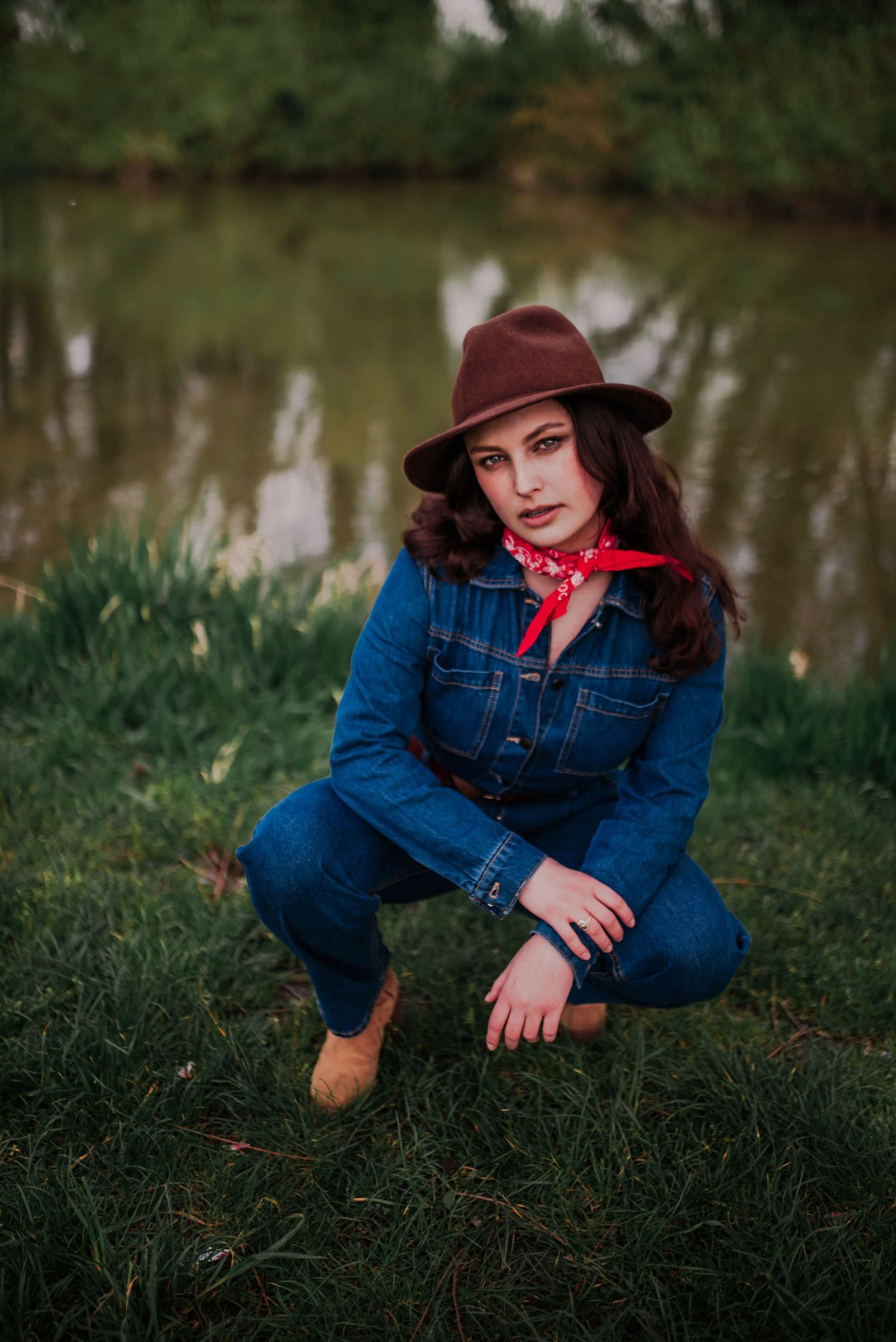 Rustic outdoor photoshoot by a pond featuring denim fashion and a burgundy hat.