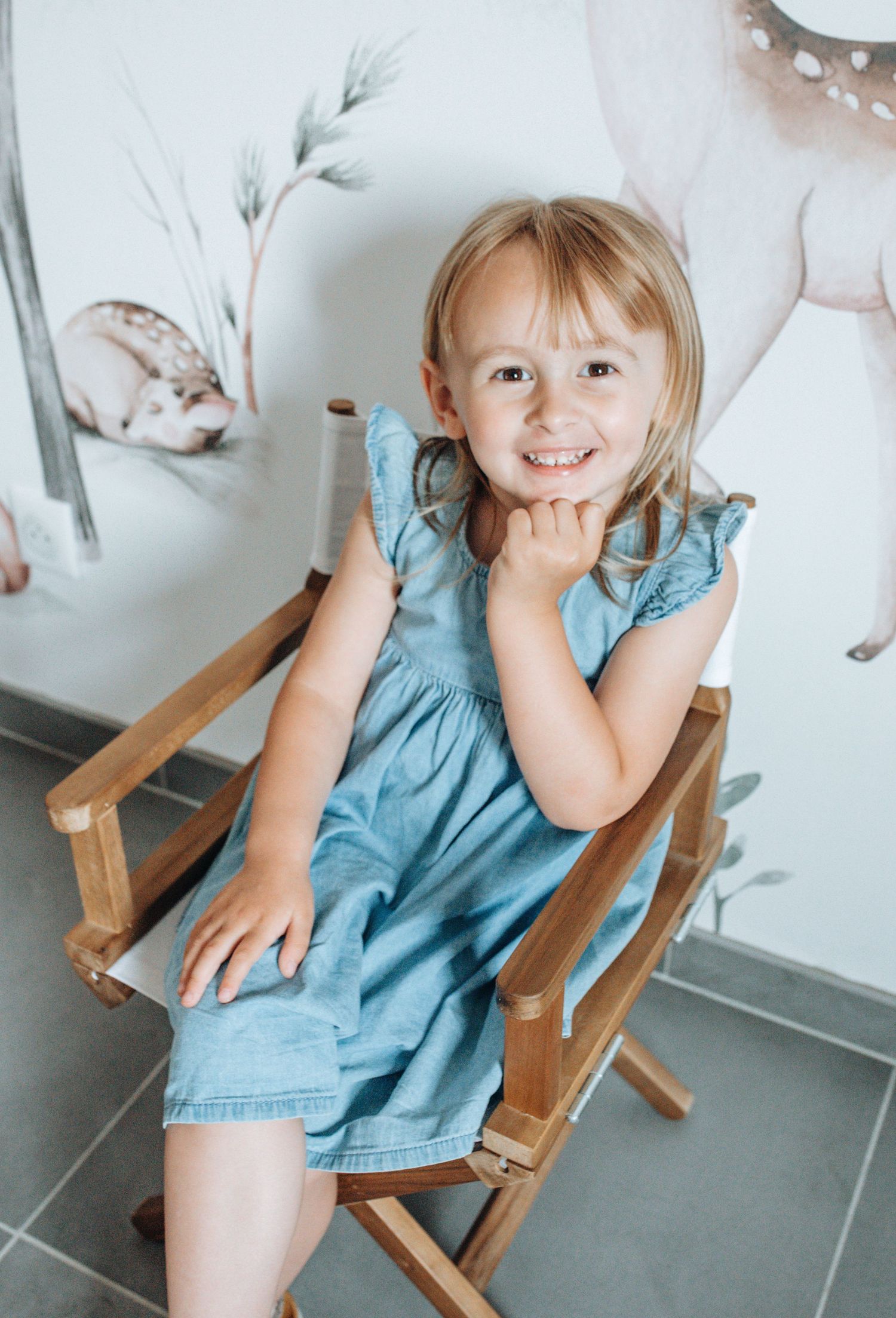 Child sitting in wooden chair with playful expressions against white wall with nature artwork.