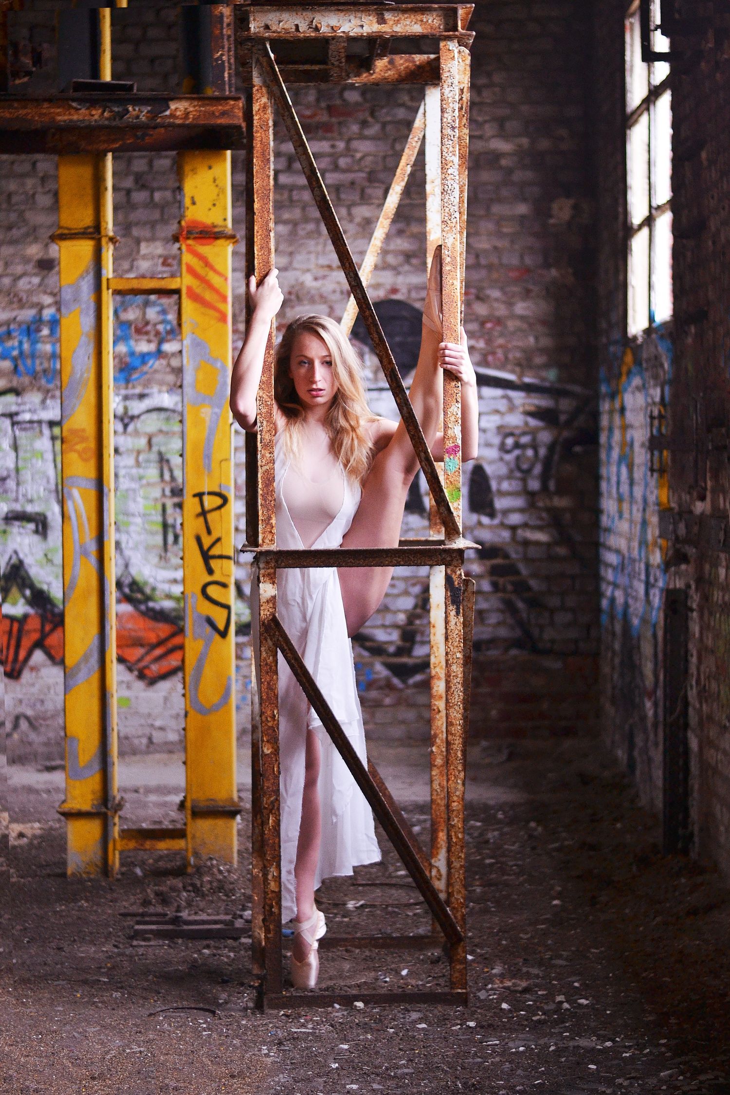 Figure in white dress poses between industrial pillars in abandoned building.