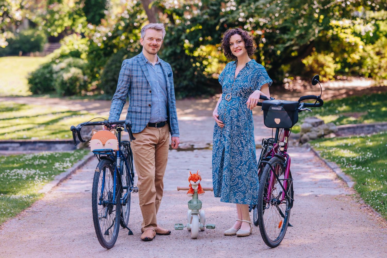 Pregnant woman and man standing with bicycles on a park path, surrounded by greenery on a sunny day.