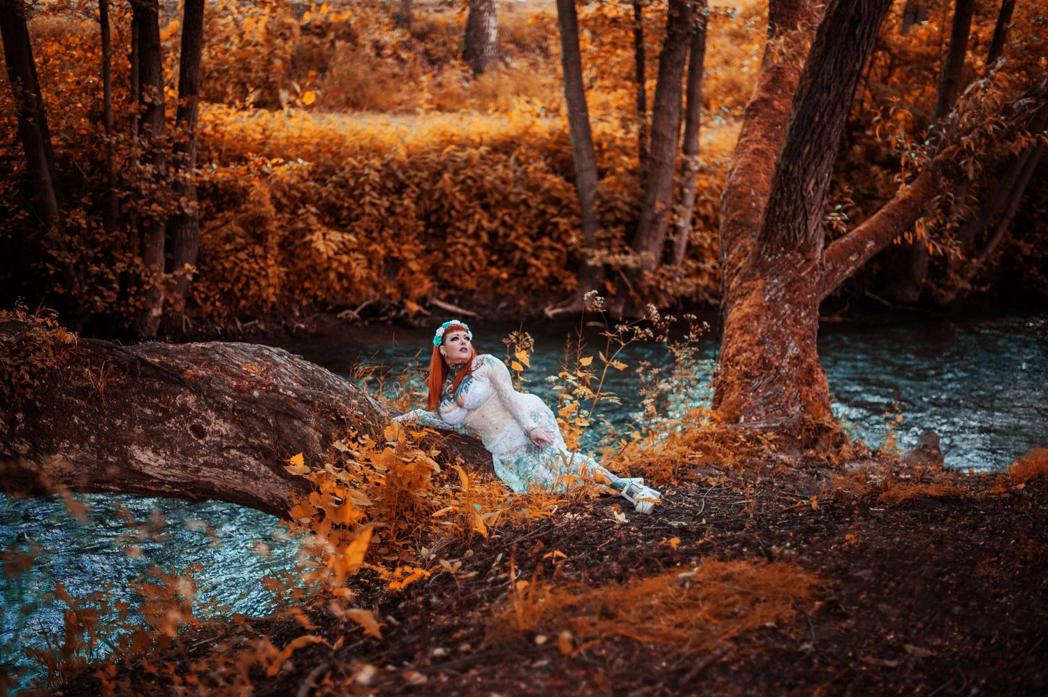 Person in flowing white dress posed by stream in autumnal forest setting.