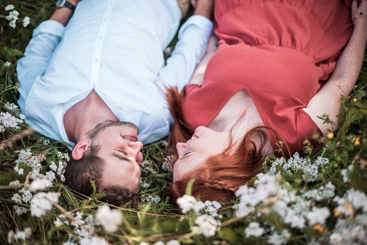 Couple lying together among white flowers in romantic outdoor photo sequence.