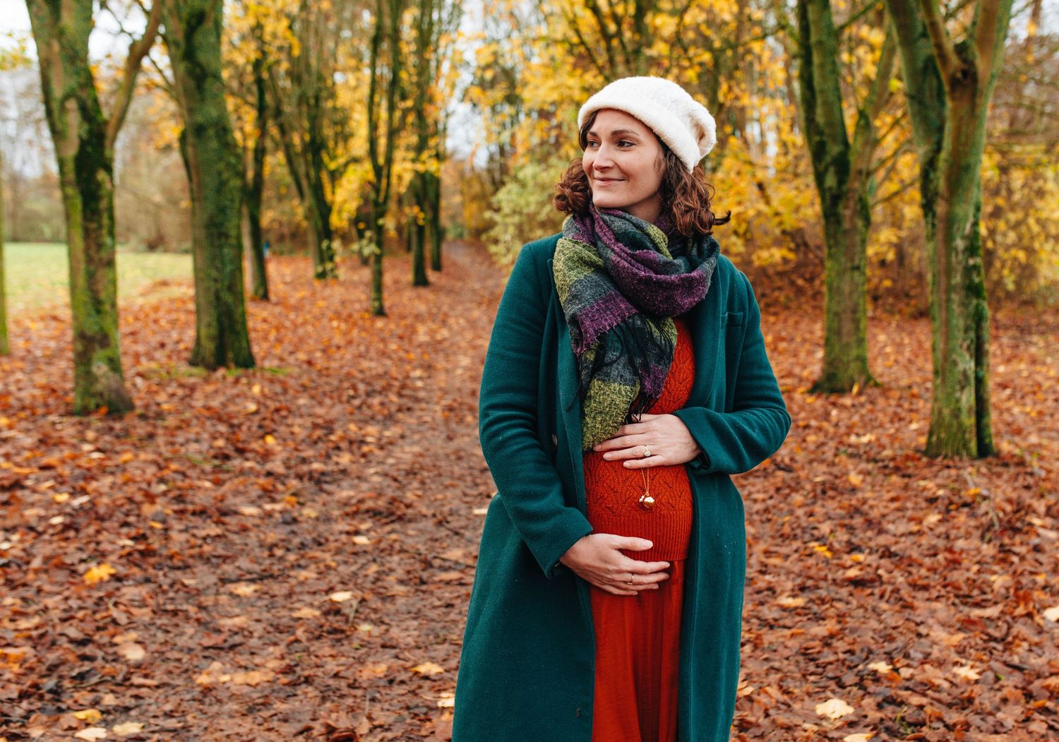 A pregnant woman in a green coat and white hat standing among autumn trees with fallen leaves.