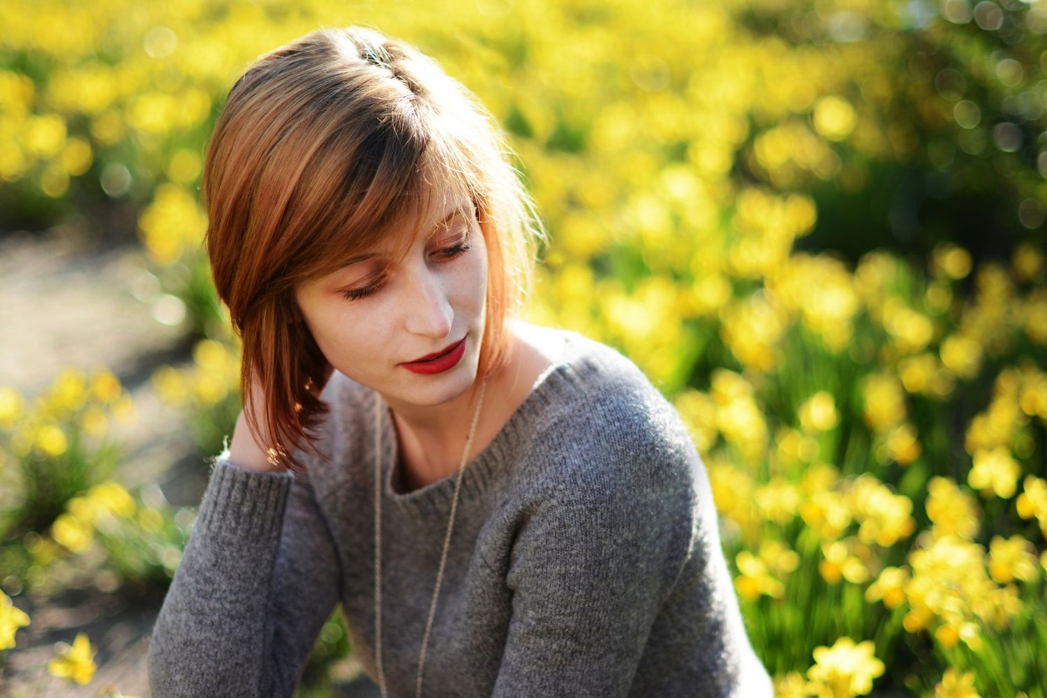 Person in gray sweater sitting outdoors among yellow flowers on sunny day.