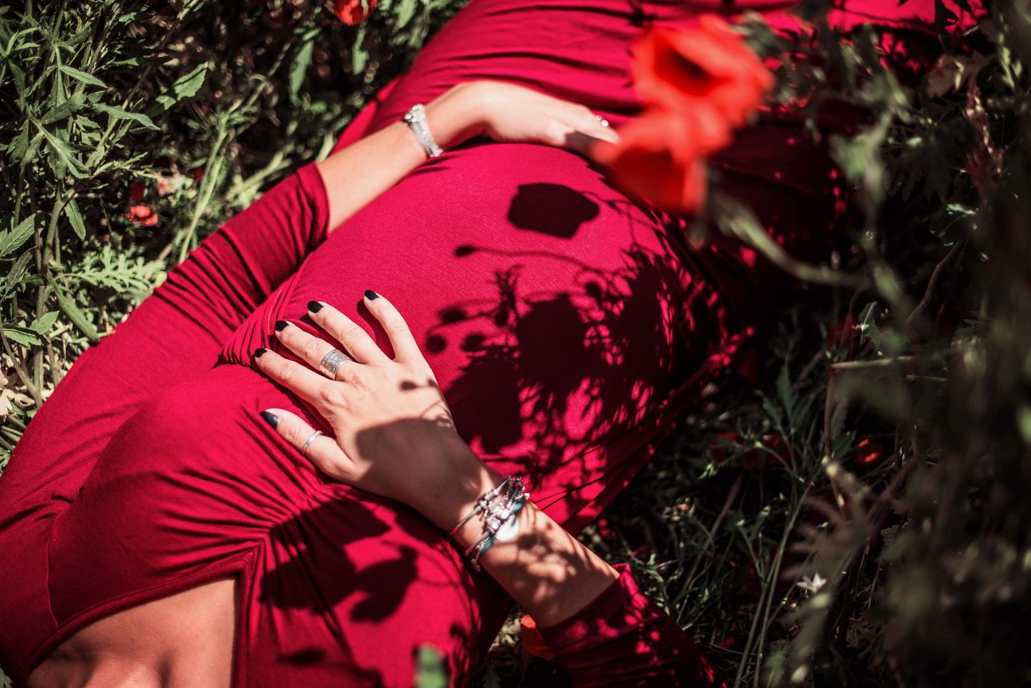 Close-up maternity photo showing hands on red dress against greenery.