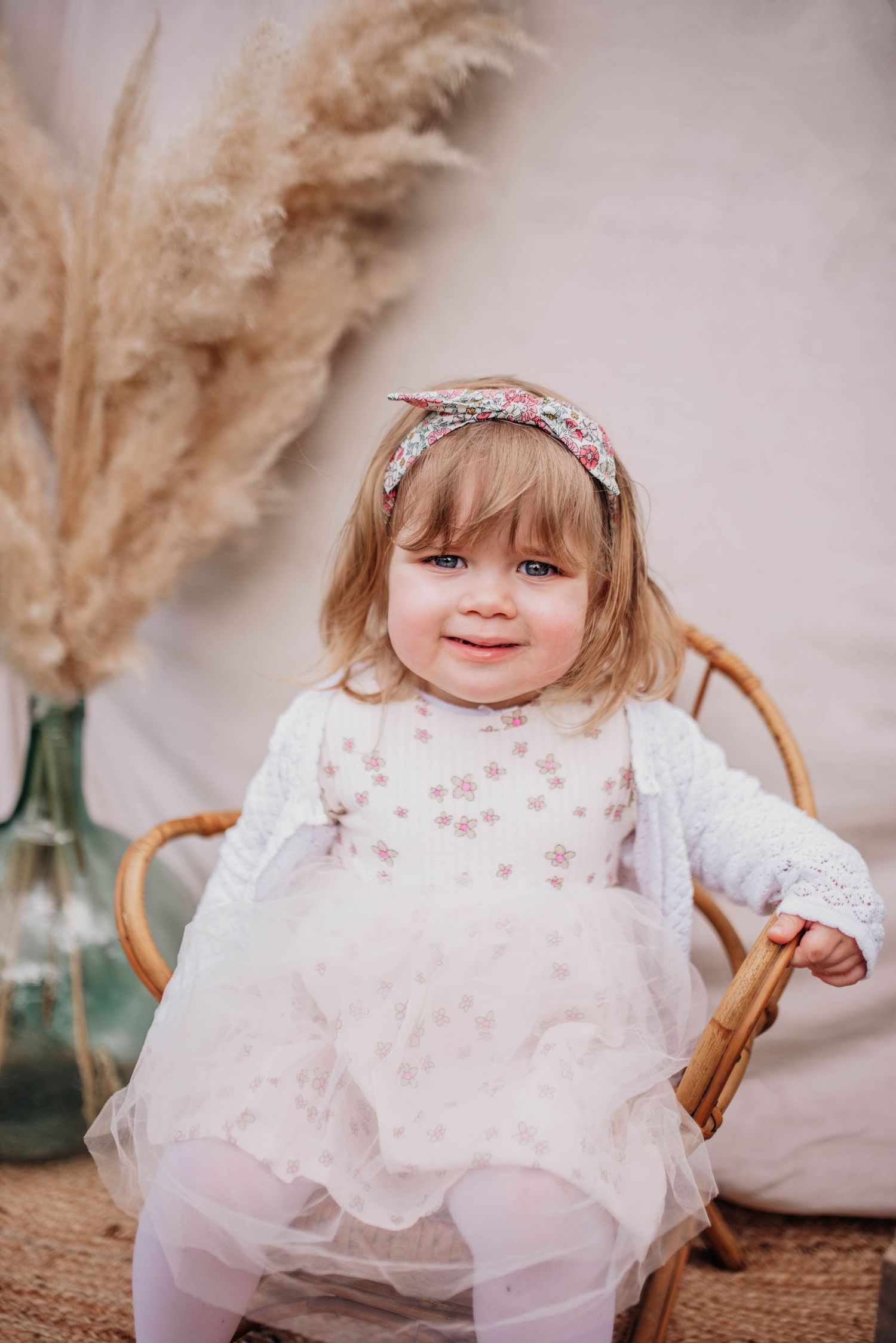Child wearing floral headband and white dress poses in vintage wooden chair.