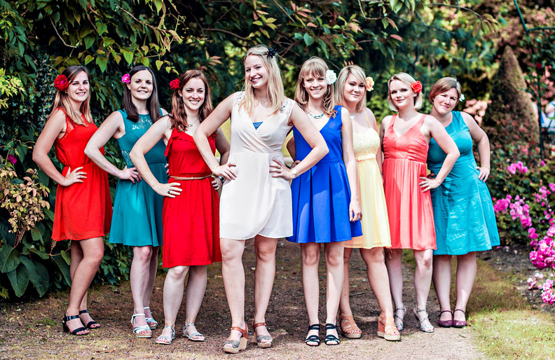 A line of friends pose in colorful summer dresses against a floral garden backdrop.