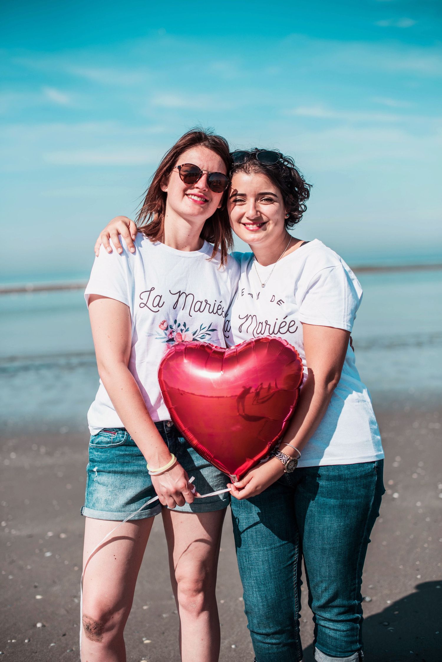 Friends posing with a red heart balloon at the beach under blue skies.