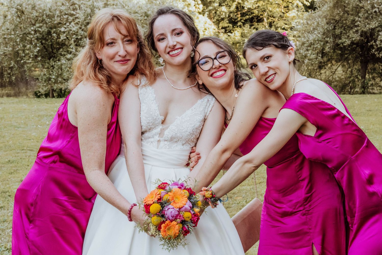 Bride in white gown holds colorful bouquet surrounded by three bridesmaids in pink dresses outdoors.