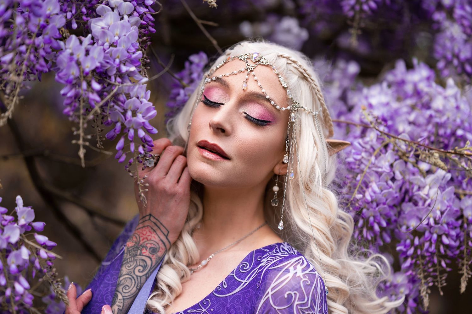 Blonde elf cosplay woman with pointed ears and crystal headpiece surrounded by purple wisteria flowers.