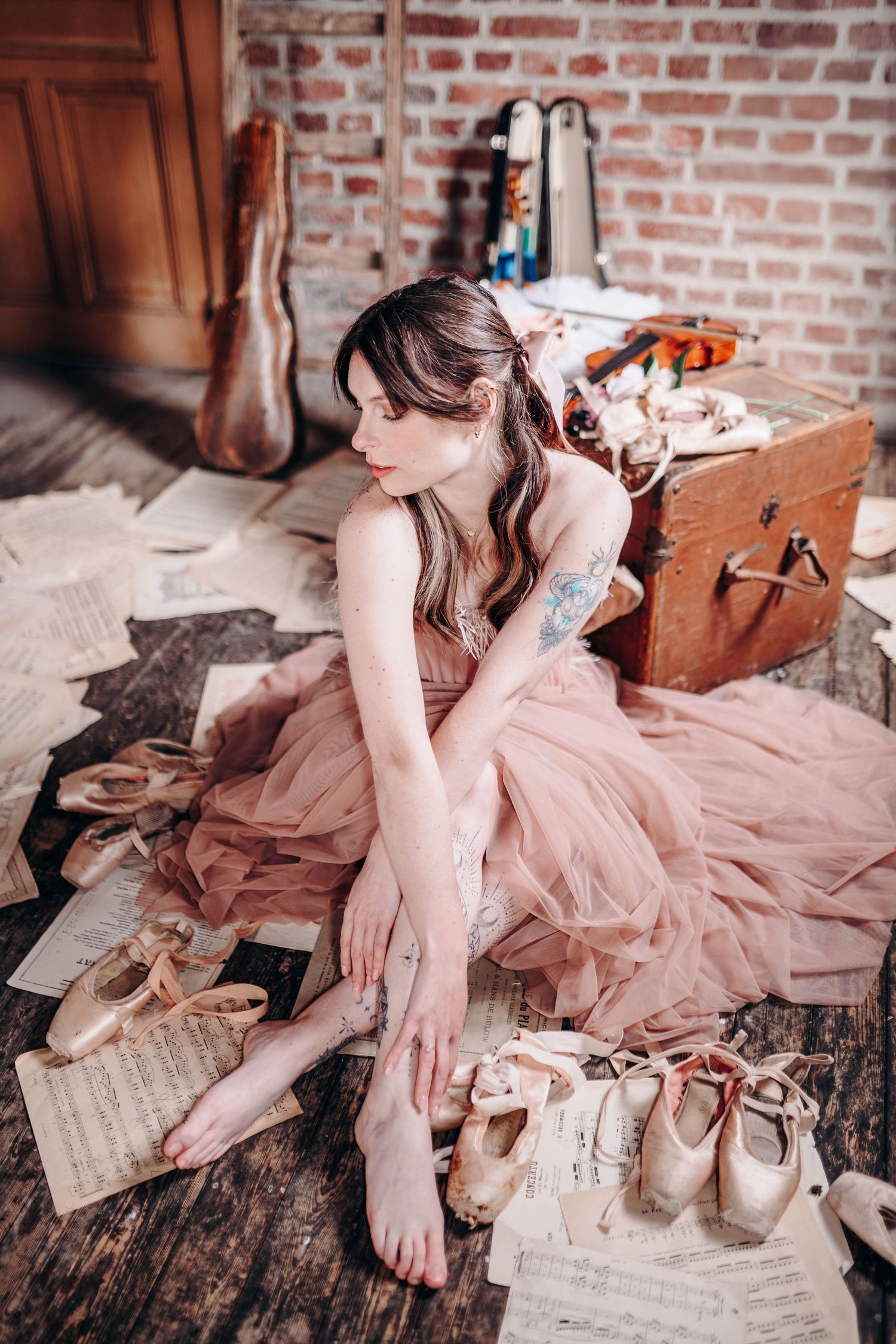 A ballerina in a pink tulle dress sits on a wooden floor surrounded by sheet music and pointe shoes near a vintage suitcase.