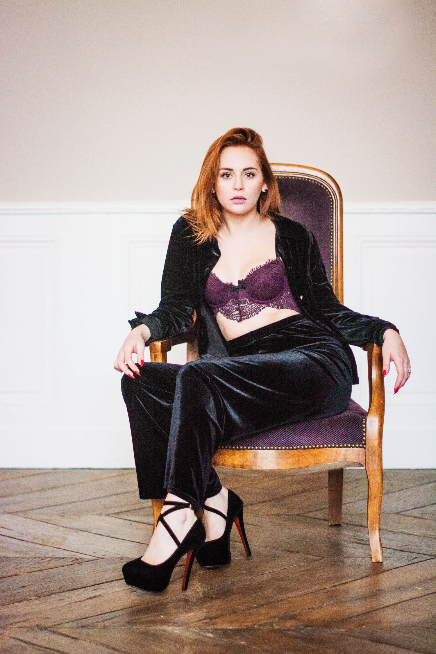 A figure in a black velvet robe and purple dress poses dramatically on a wooden chair against a light background.
